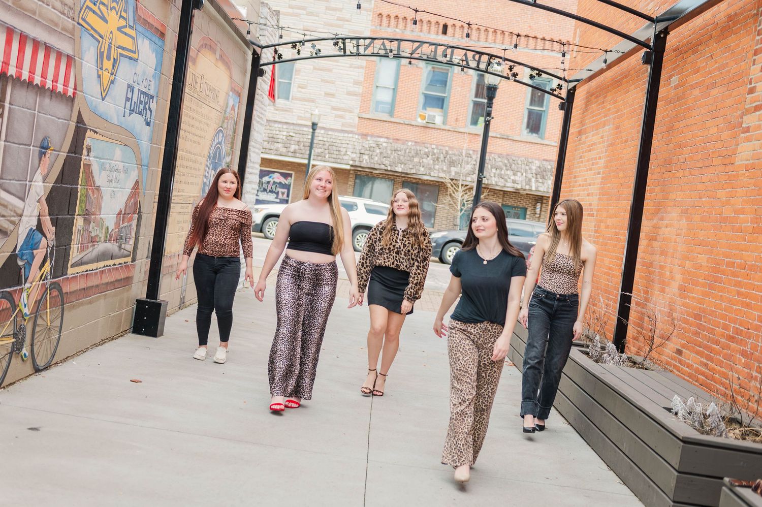 Five young women walk confidently down an urban alley with brick walls and colorful murals on a sunny day.