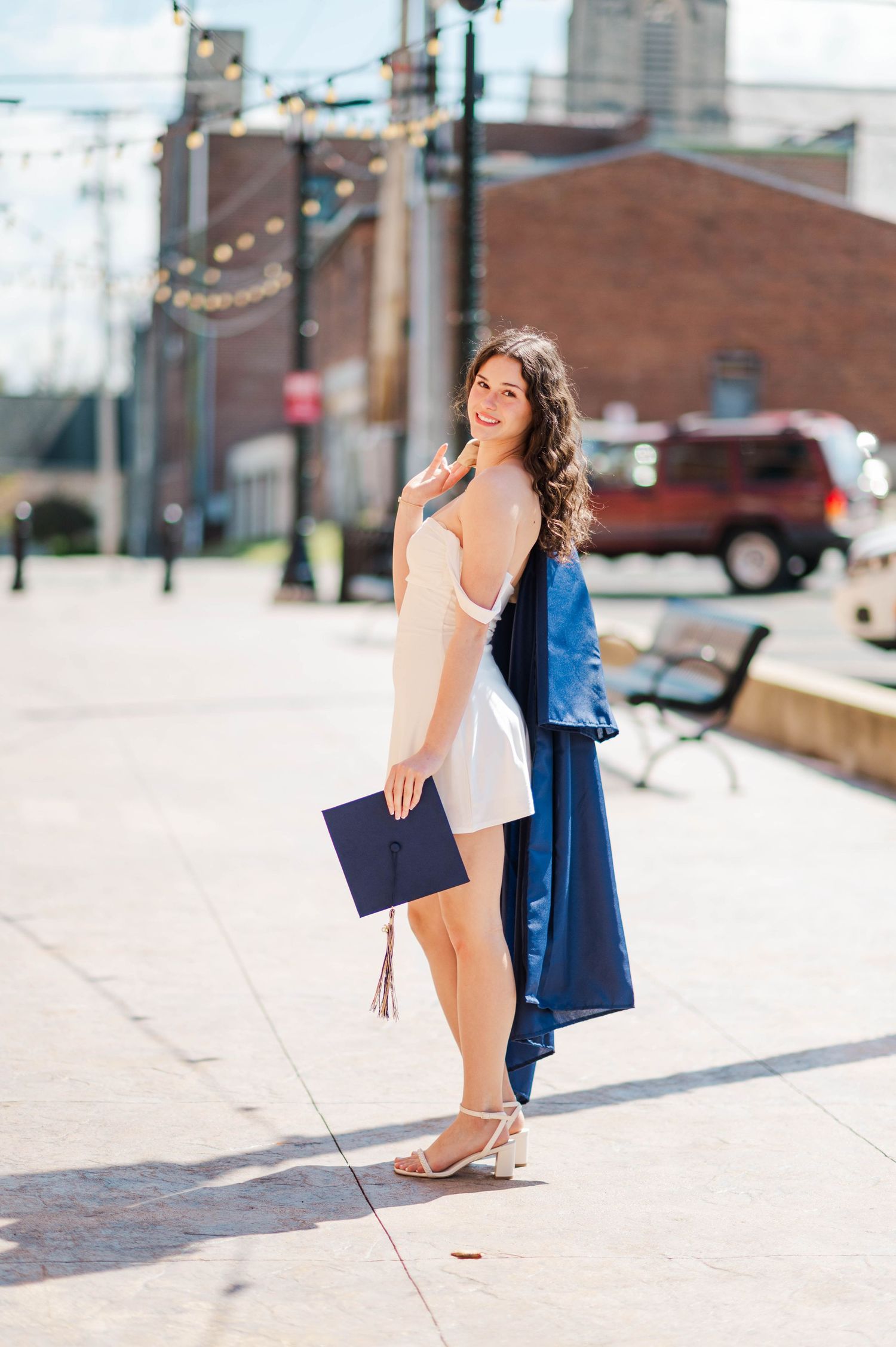 Young woman in white dress holding navy graduation cap on sunny street.