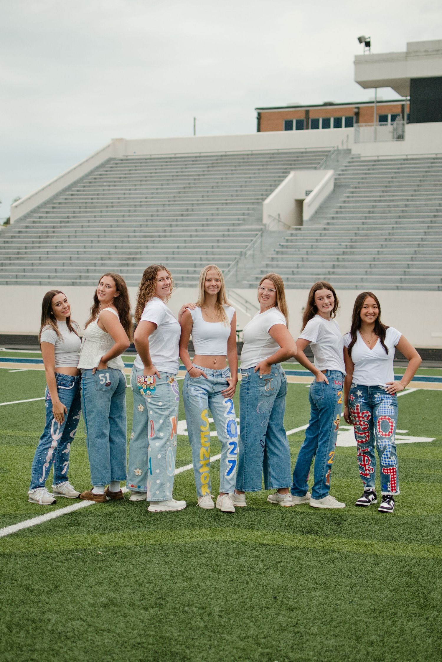 Seven teenage girls in white tops and decorated denim jeans pose together on a football field with stadium bleachers behind them.