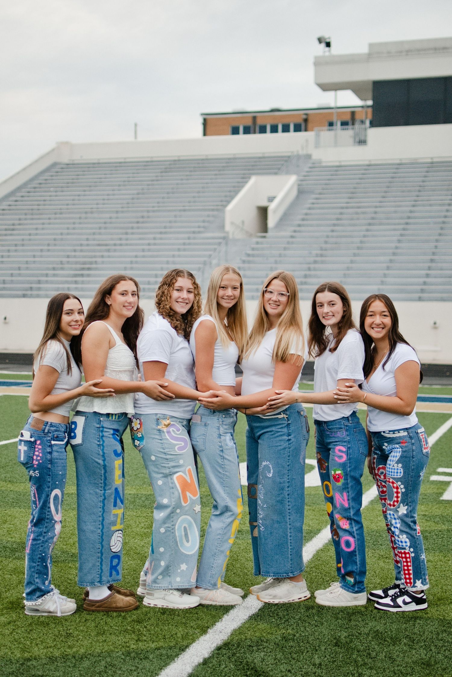 Seven teen girls in white tees and decorated senior jeans pose together smiling on a football field.