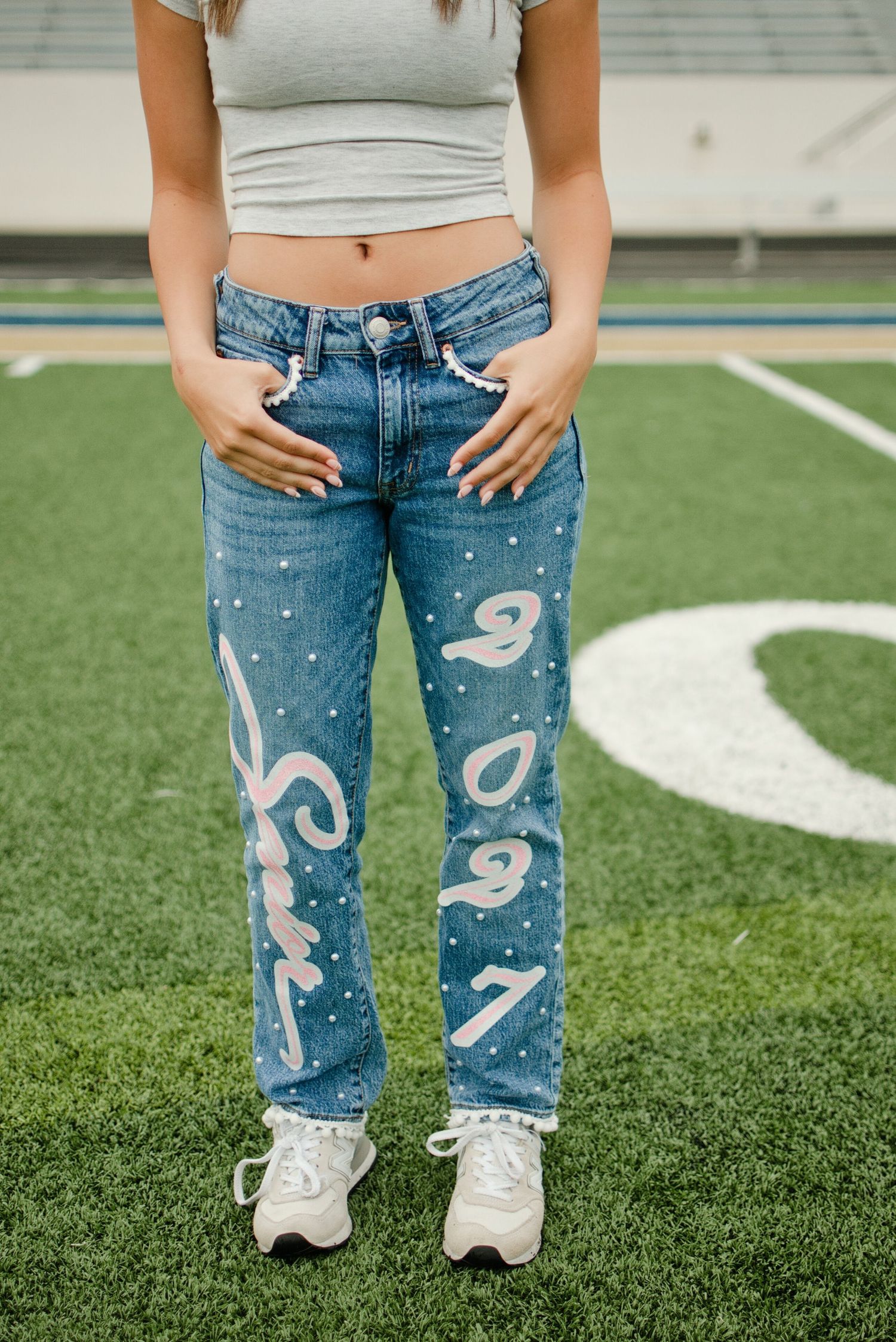 Close-up of decorated denim jeans with painted script lettering and pearl studs worn on a green football field turf.