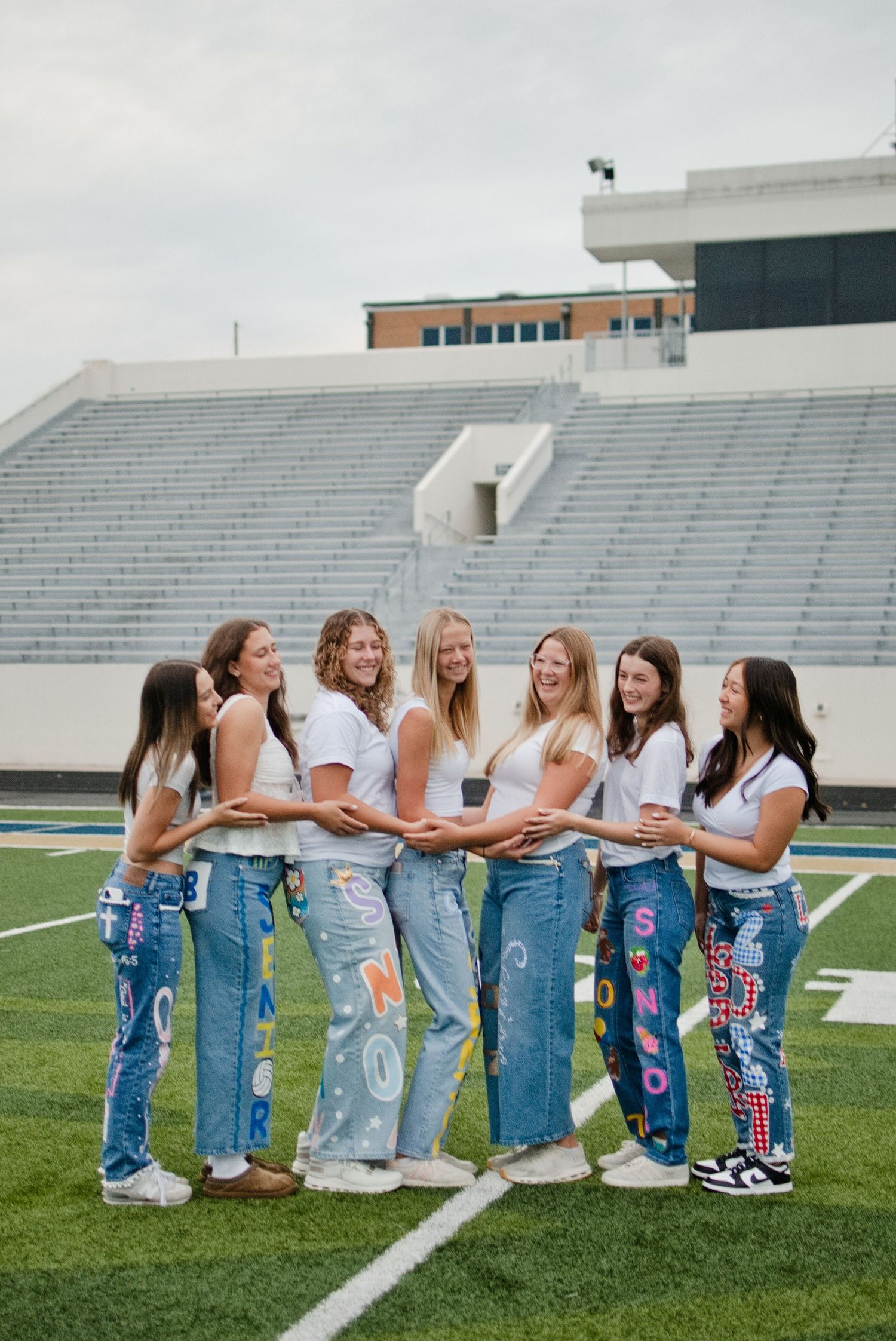 Seven girls in white tops and painted senior 2027 jeans pose together smiling on a football field.