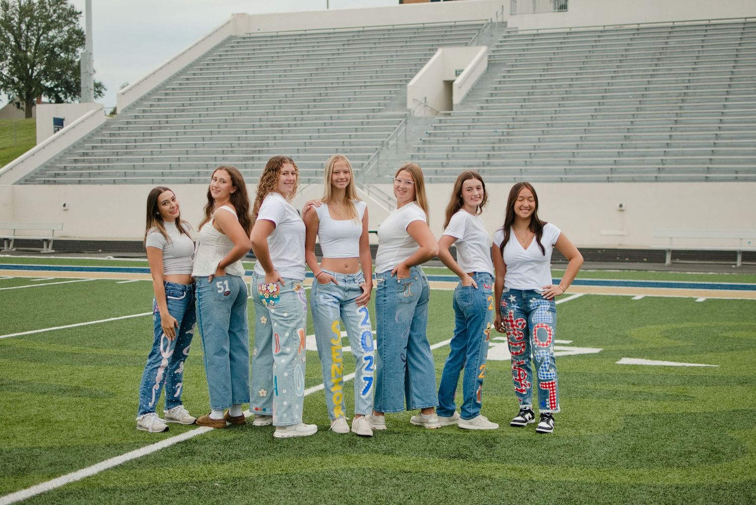 Seven young women in white tops and jeans pose together on a football field in front of stadium bleachers.