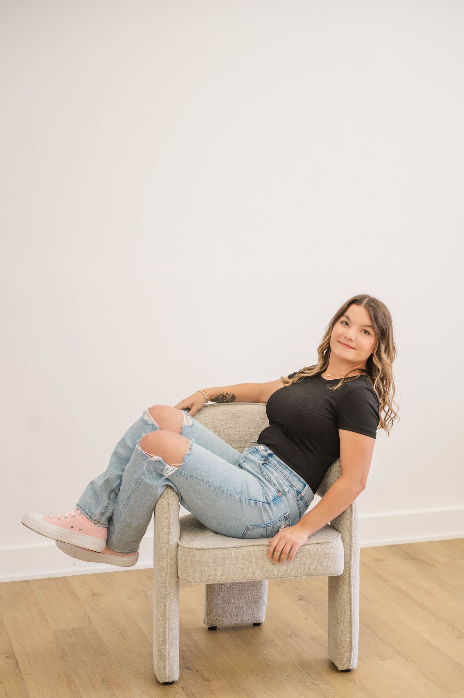 Young woman in black tee and ripped jeans reclines in modern chair wearing pink sneakers, posing casually.