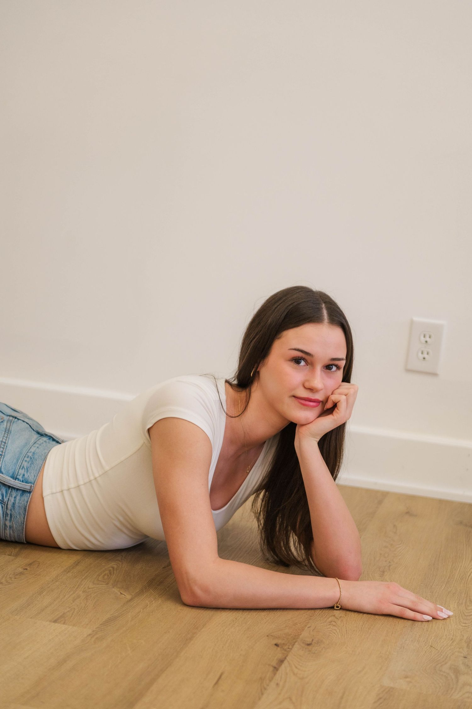 Young woman in white top and denim shorts lying on hardwood floor, resting chin on hand in a relaxed pose.