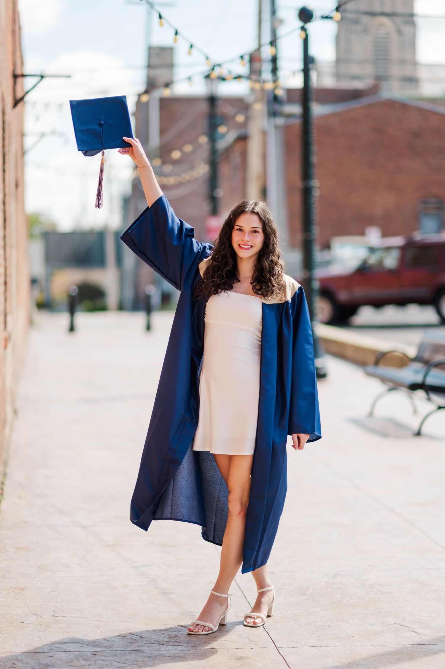 Joyful brunette graduate in navy gown raising mortarboard cap while walking on city sidewalk.