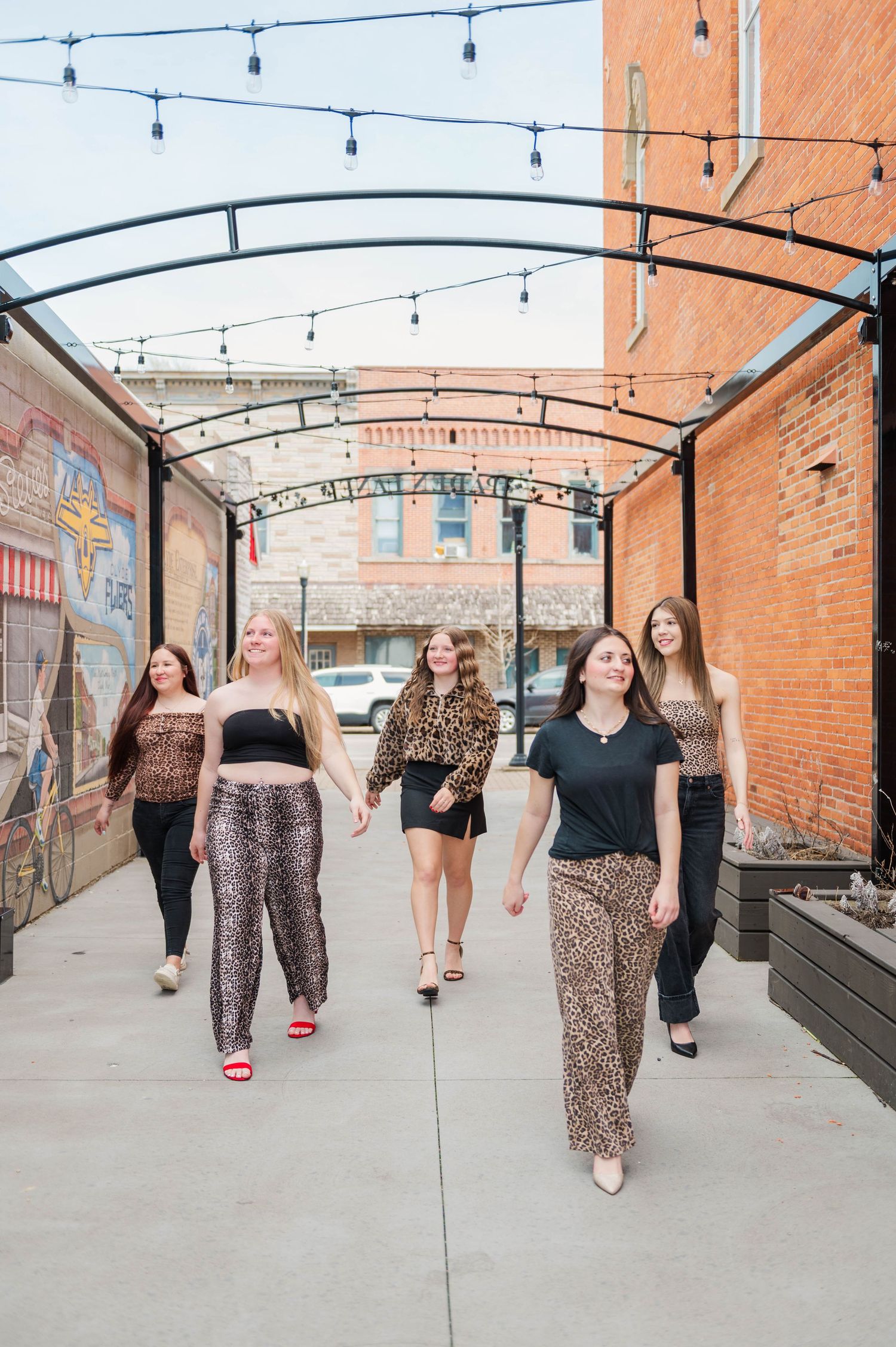 Five young women walk together under string lights in coordinating animal print outfits in urban alley.