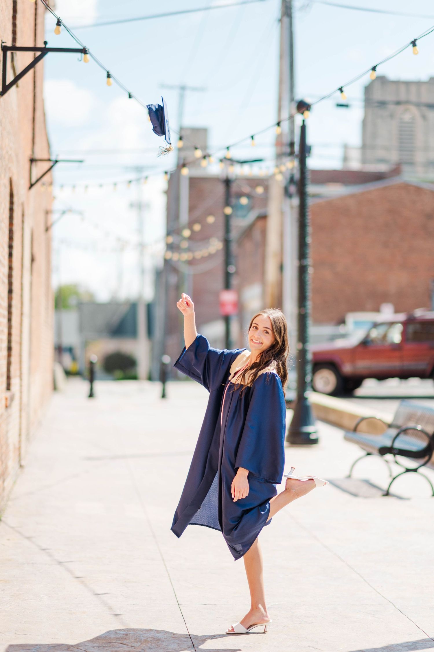Graduate in navy gown playfully tosses her cap on a sunlit brick-lined city sidewalk.
