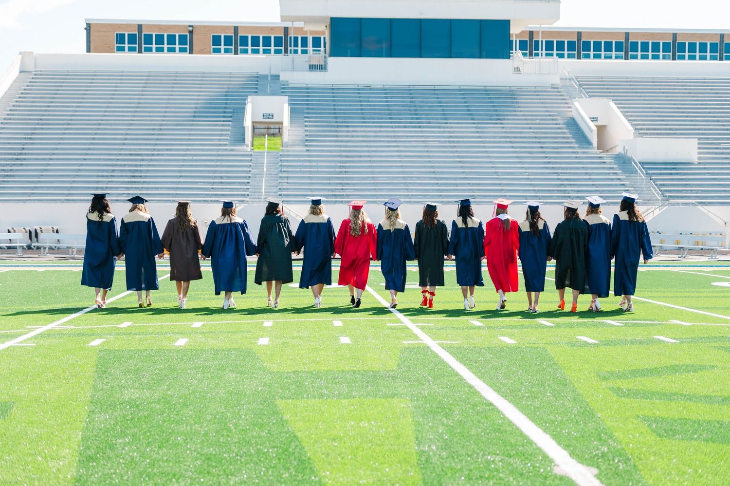 A group of graduates in caps and gowns stand together on a football field stadium, viewed from behind.