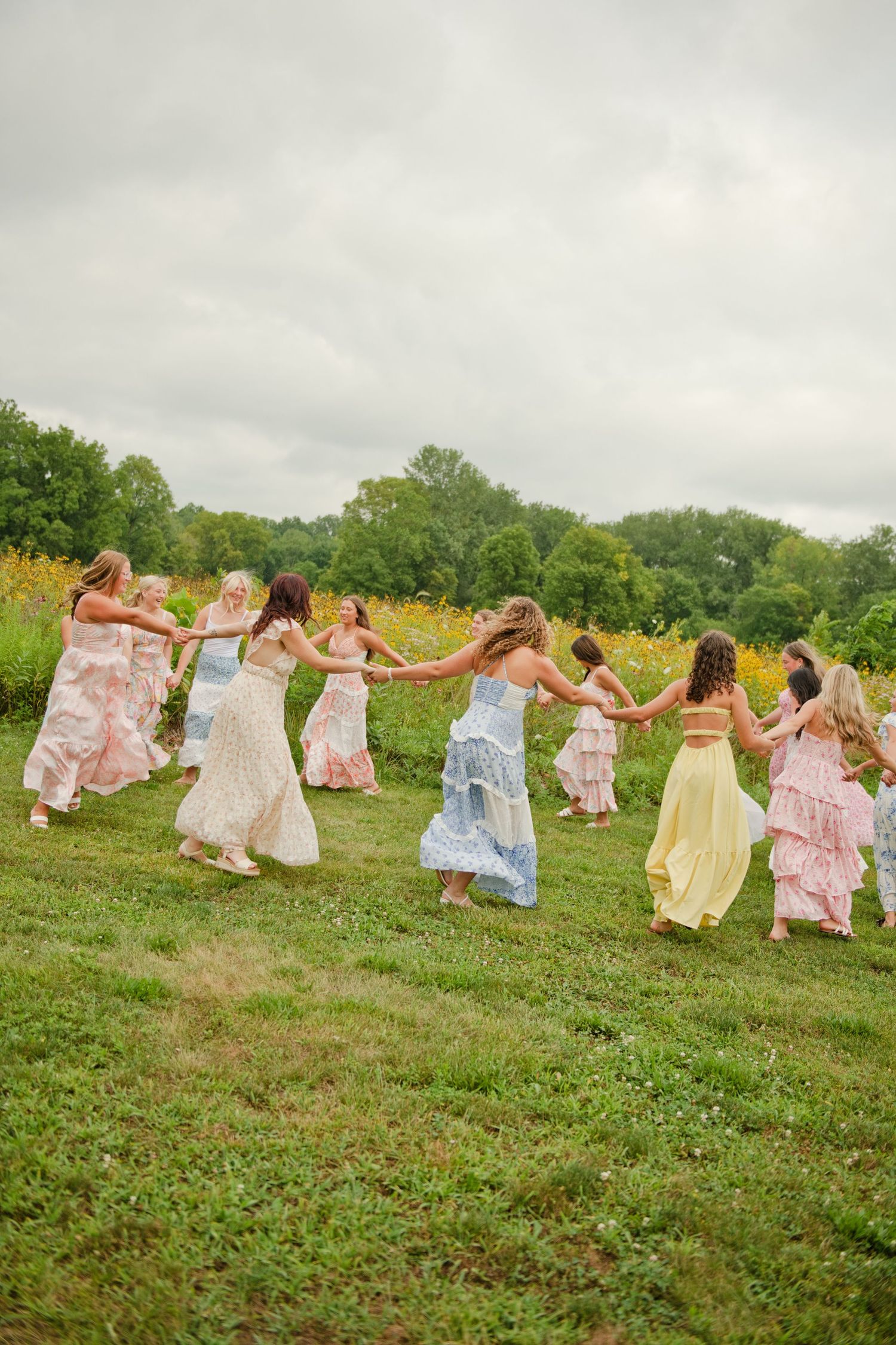 A group of women in pastel maxi dresses hold hands in a line, dancing on a green meadow with trees in the background.