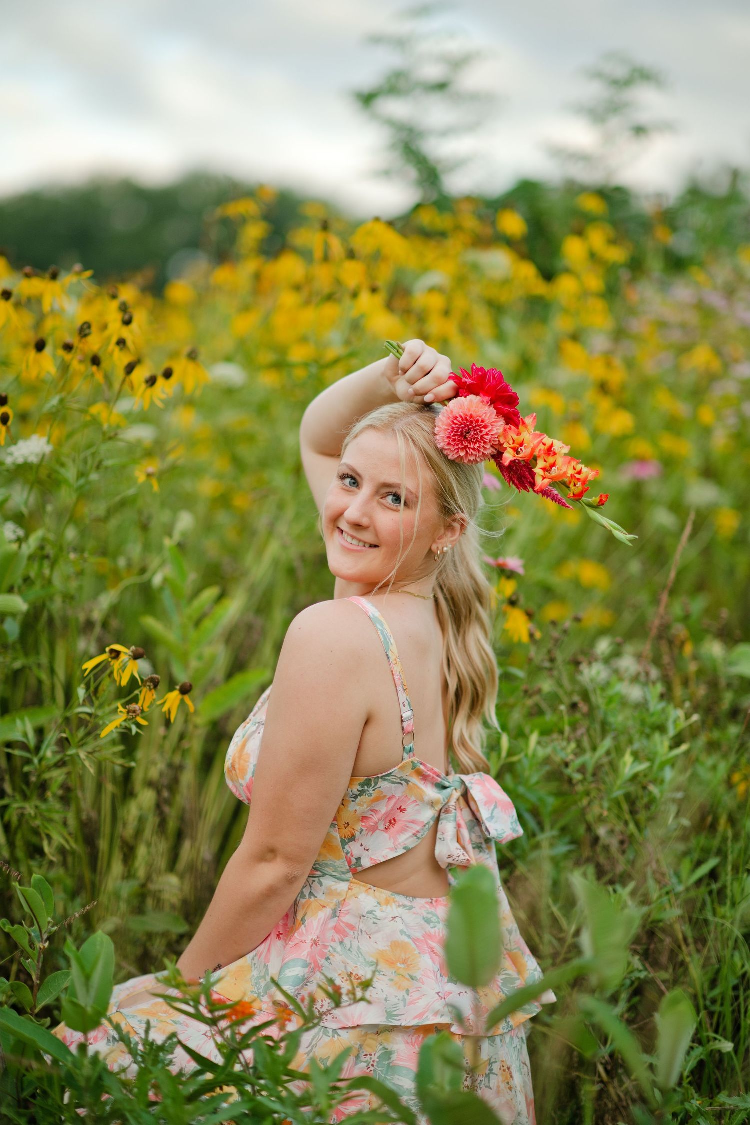 Smiling blonde woman in floral dress holding flowers in a yellow wildflower field.