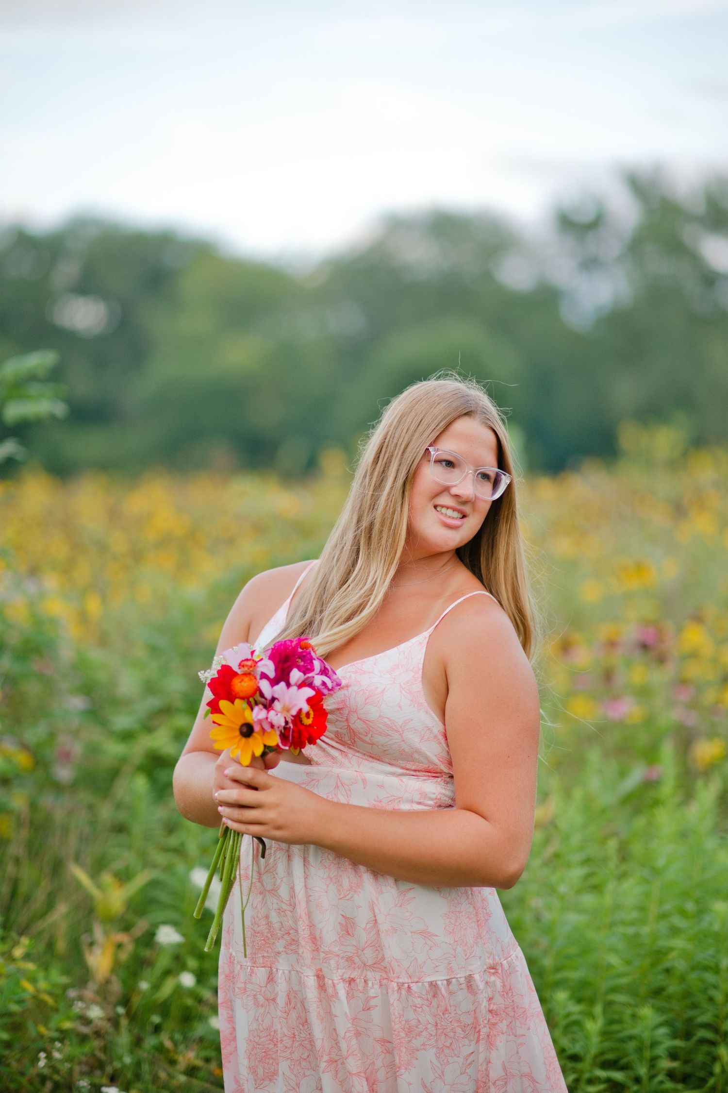 Smiling blonde woman in pink floral dress holding colorful bouquet in a wildflower field.