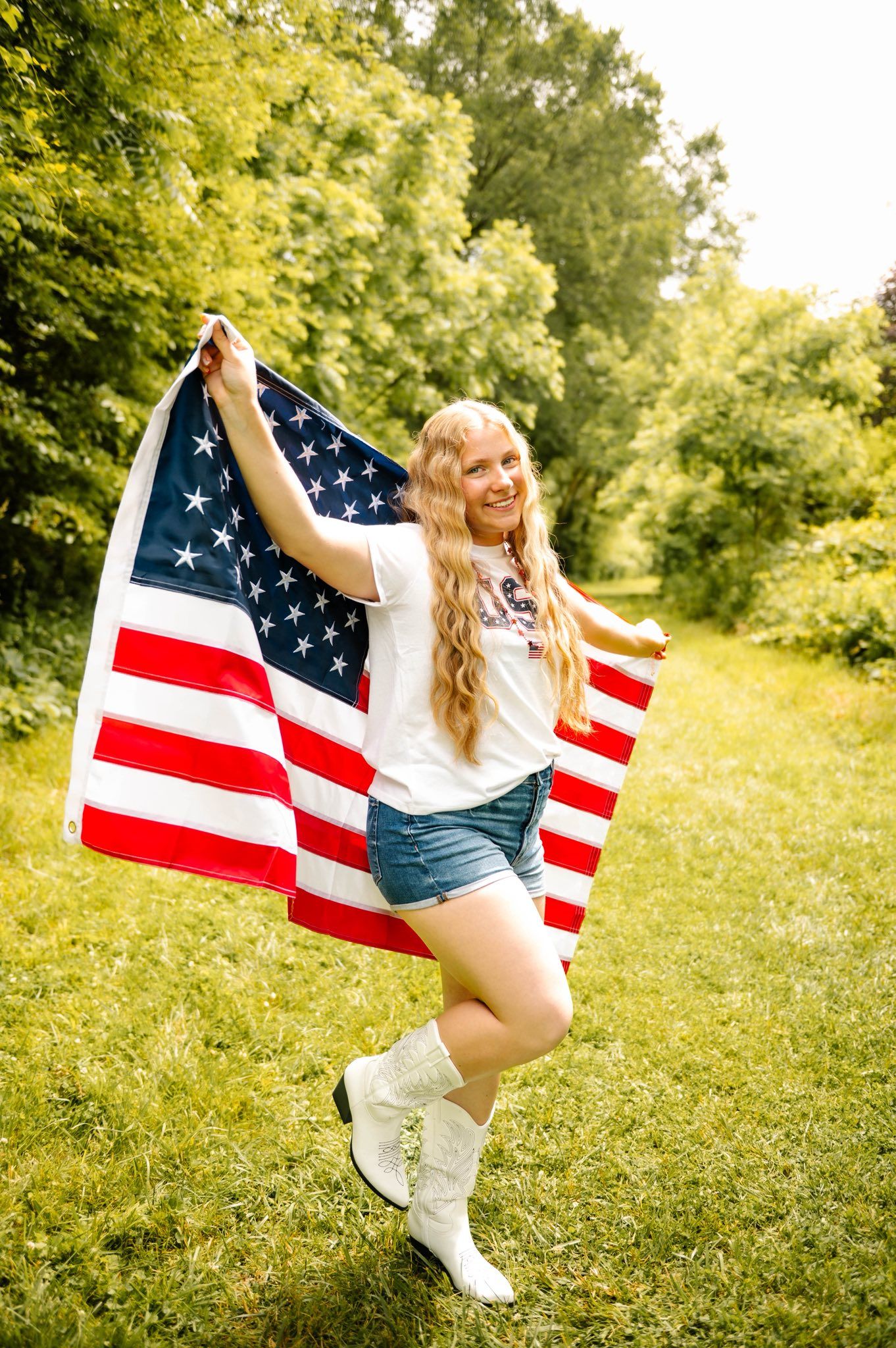 Someone poses with an American flag in a sunny field wearing casual summer attire.