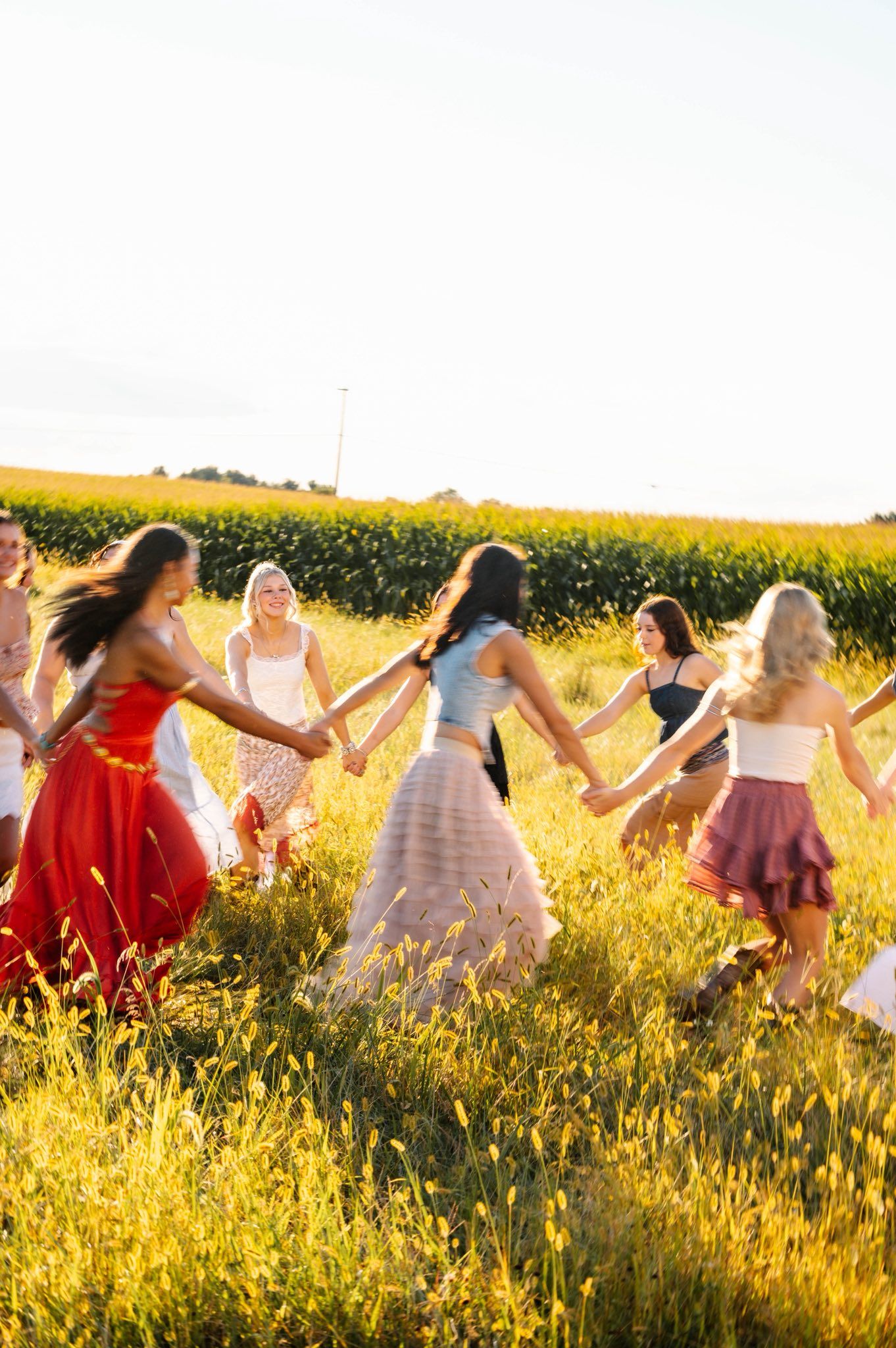 People holding hands and dancing in a line through a sun-drenched field of yellow flowers.