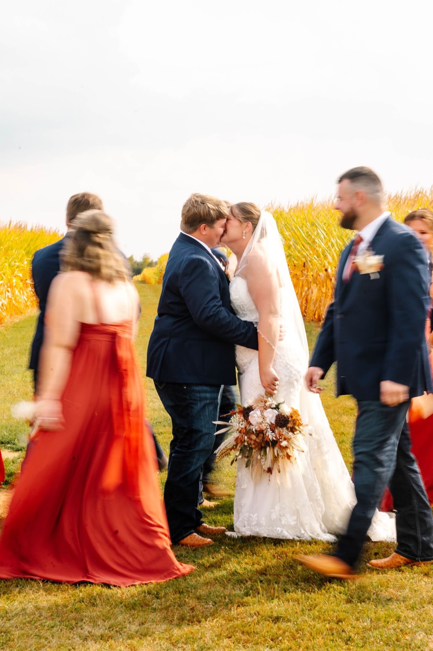 Wedding party poses in a golden wheat field with bridesmaids in red dresses and groomsmen in navy suits at sunset.
