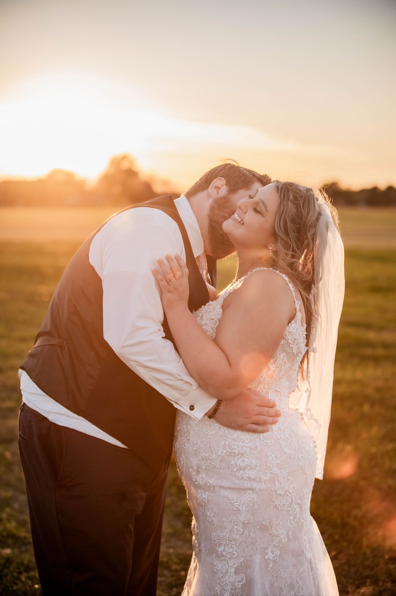 Newlyweds embrace during sunset in an open field wearing formal wedding attire.