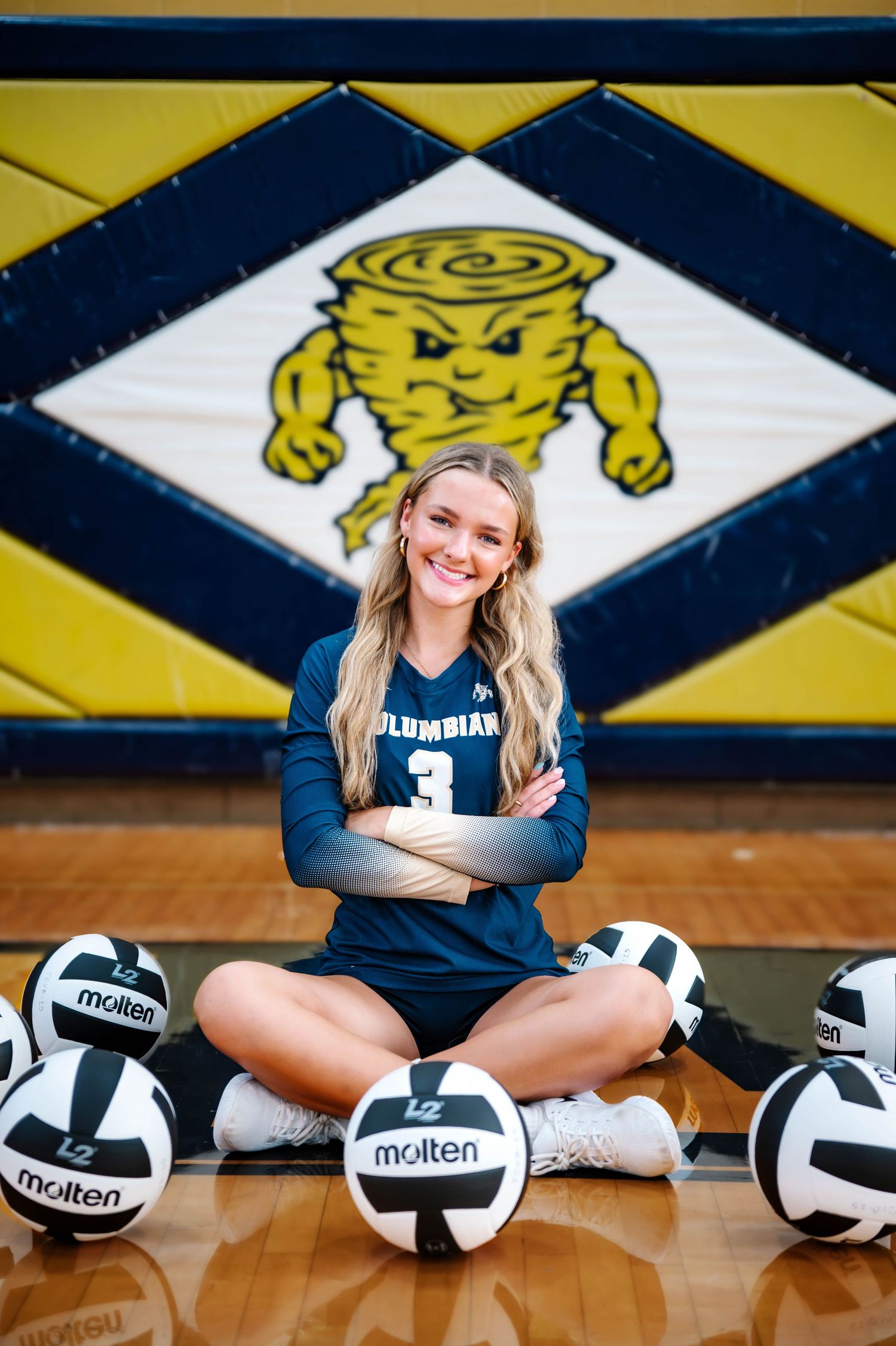 Volleyball player poses with Molten volleyballs in a school gym with mascot artwork.