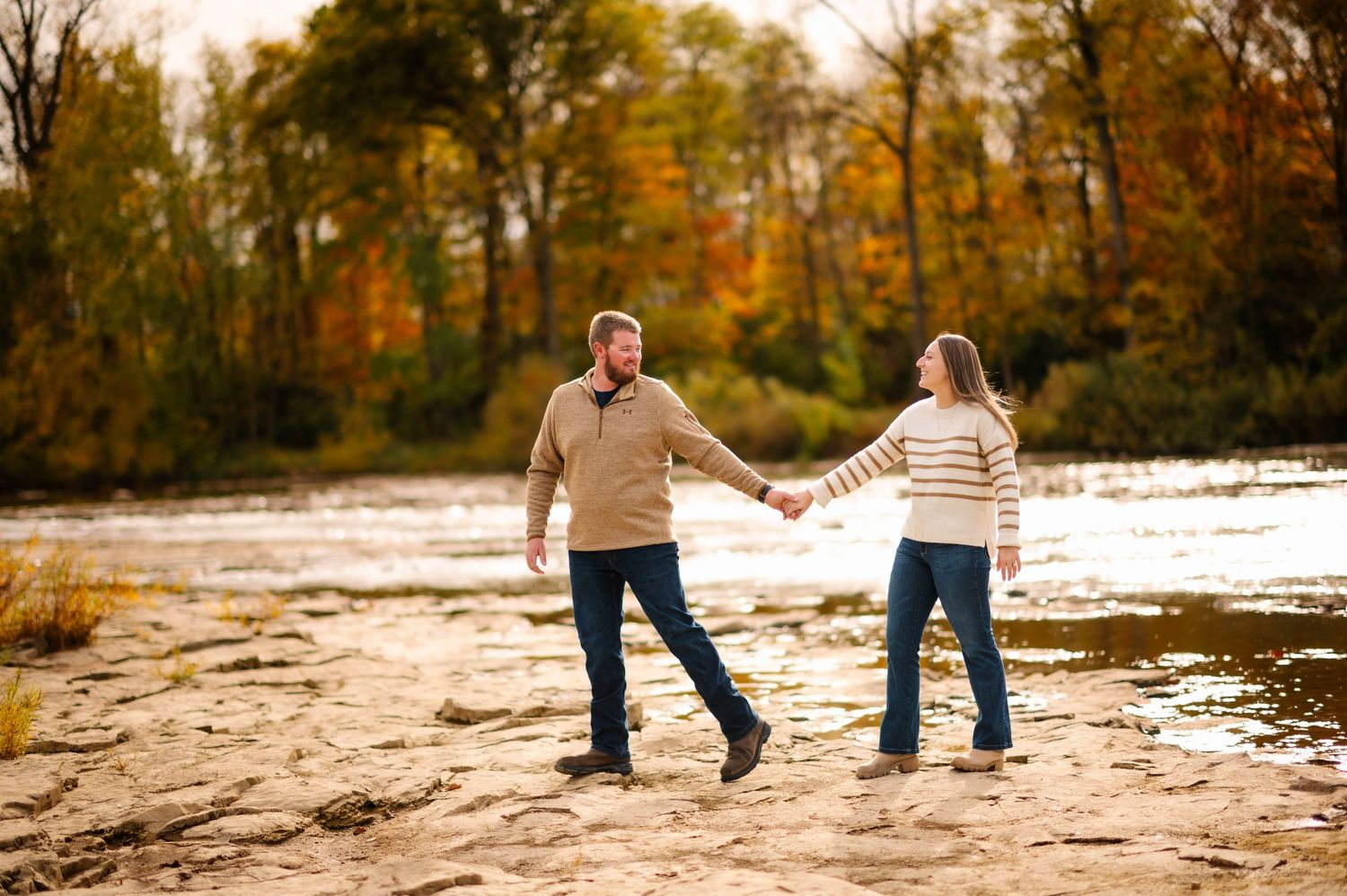 Couple walking hand in hand along rocky shoreline with autumn trees in background.