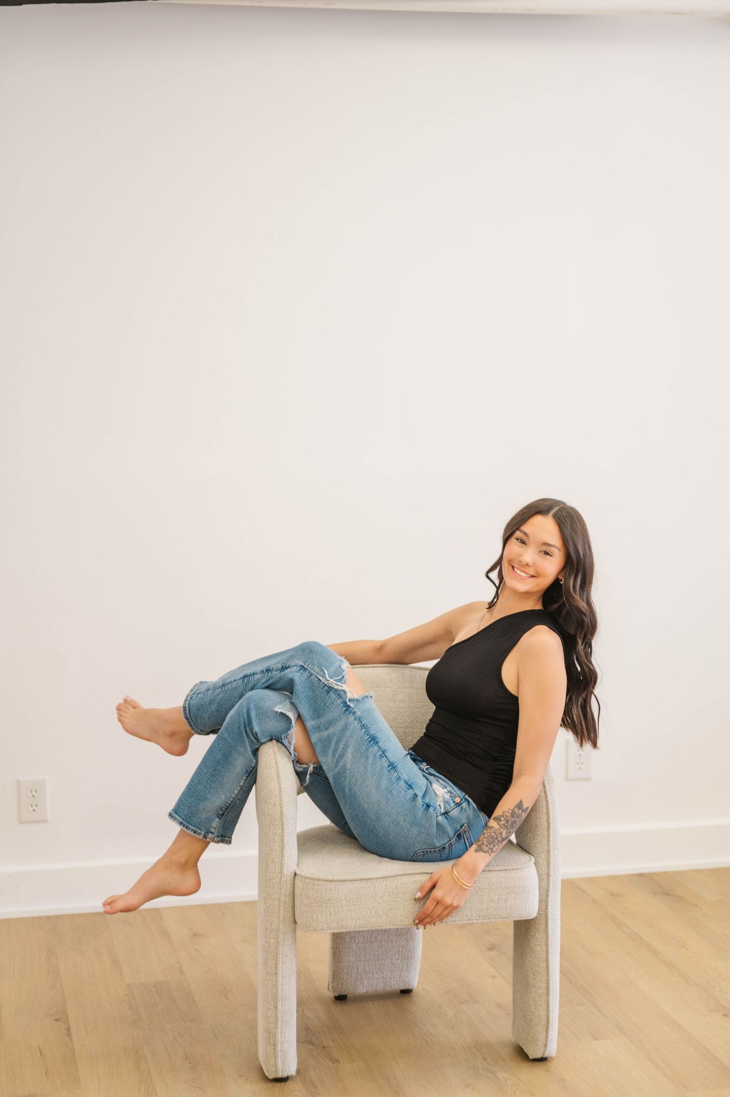 Woman in black top and ripped jeans sitting relaxed in modern chair, smiling at camera.