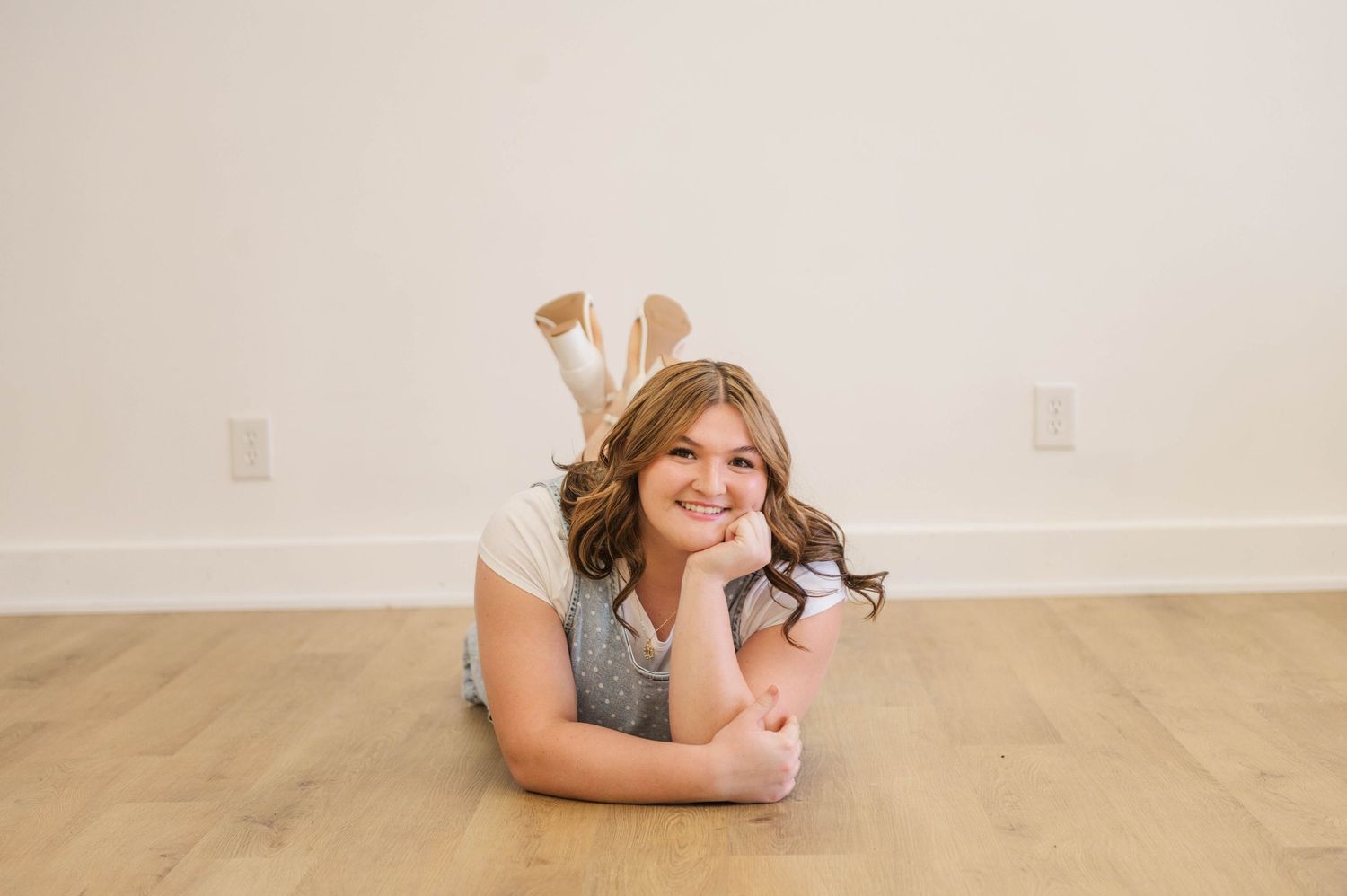 Young girl in gray outfit lying on hardwood floor with chin resting on hands, smiling.