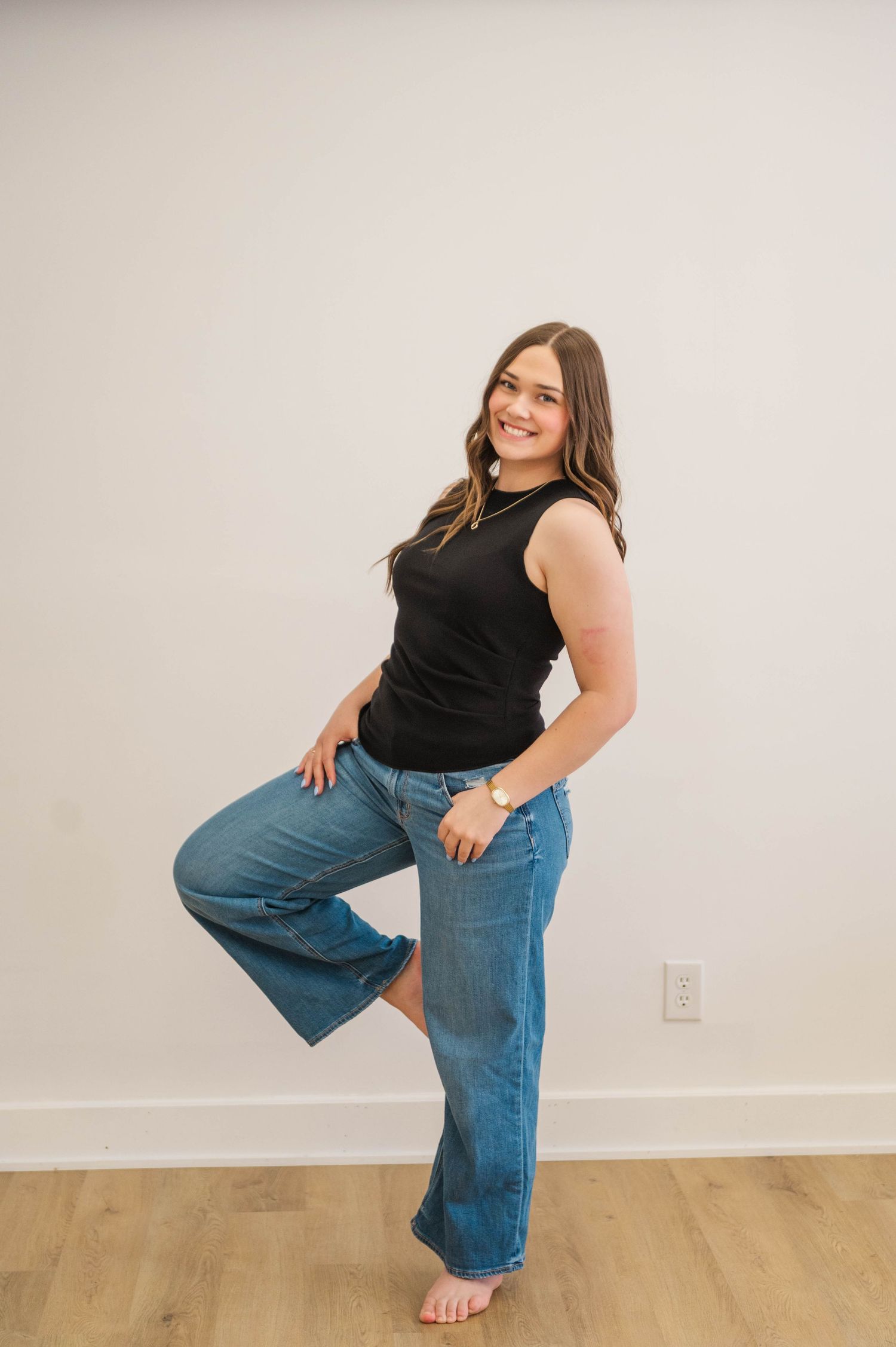 Woman in black sleeveless top and wide-leg jeans posing barefoot on hardwood floor.