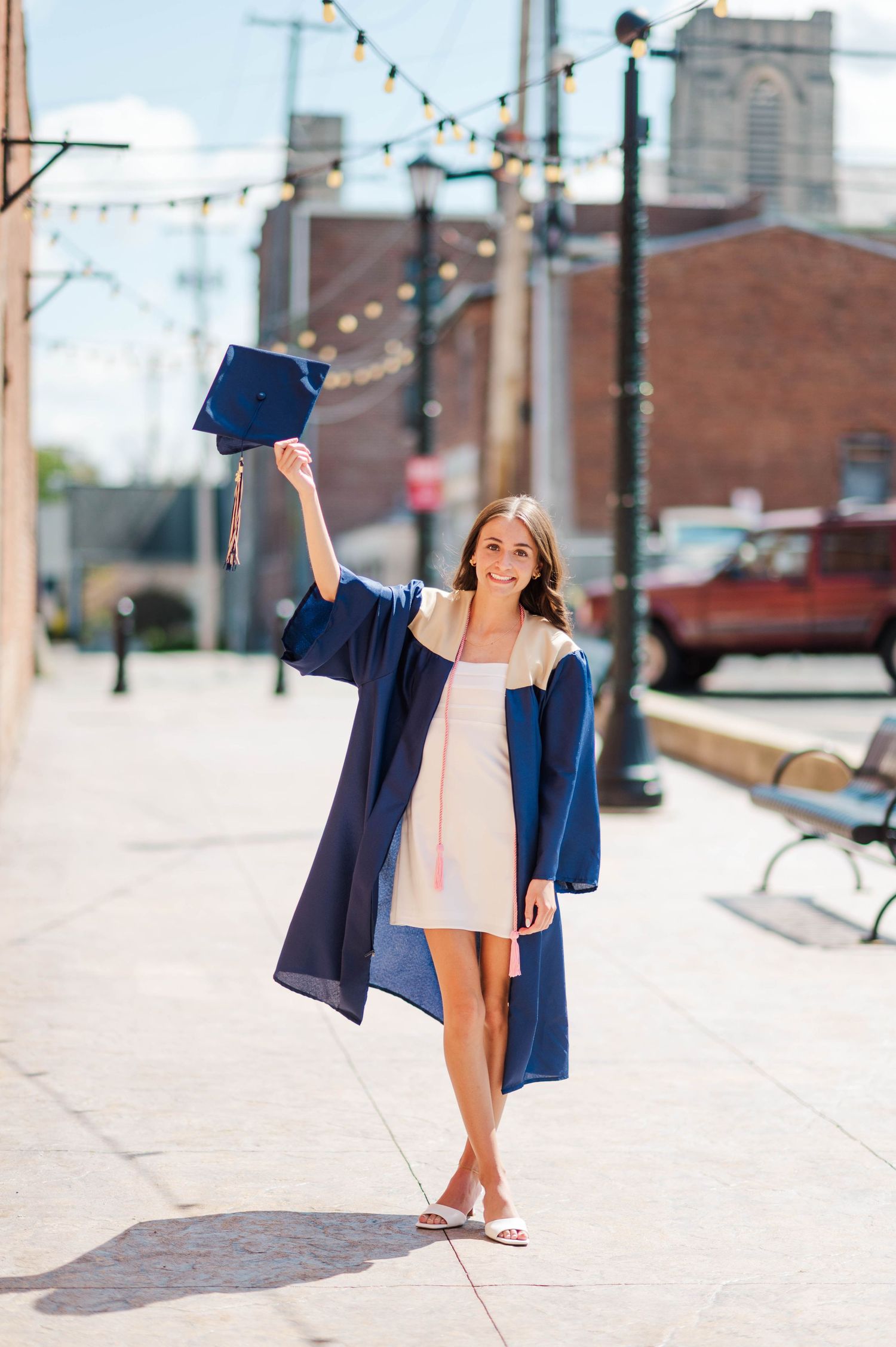Joyful graduate in navy gown holding cap up on sunny brick-lined sidewalk.