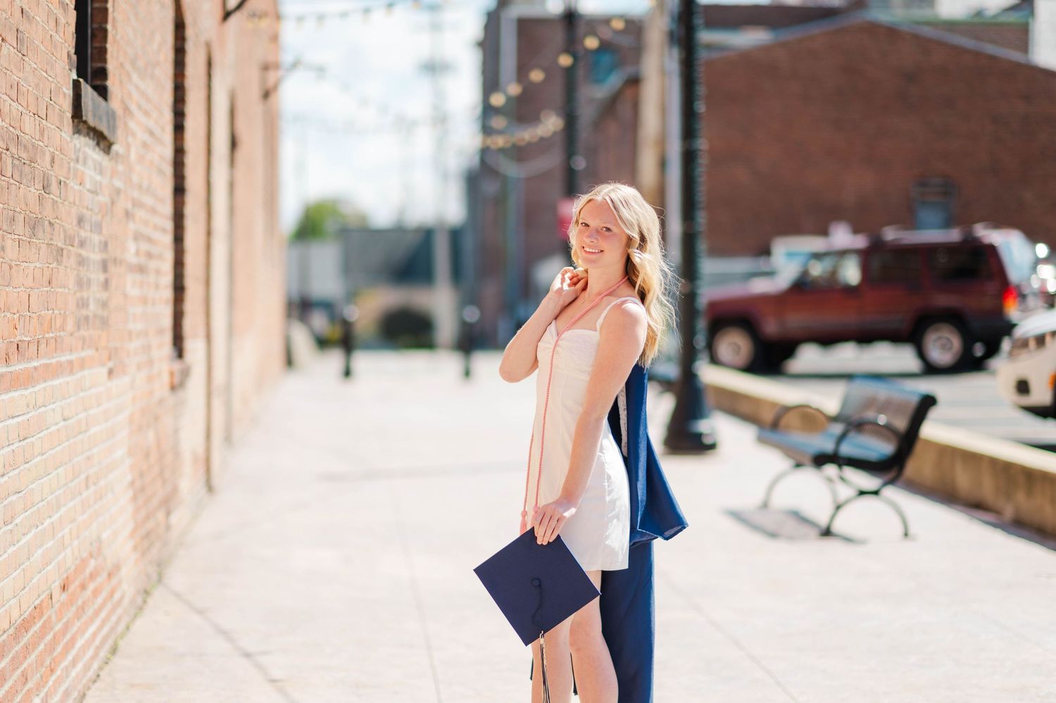 Blonde woman in white dress holding navy graduation cap while standing in sunny urban alley.