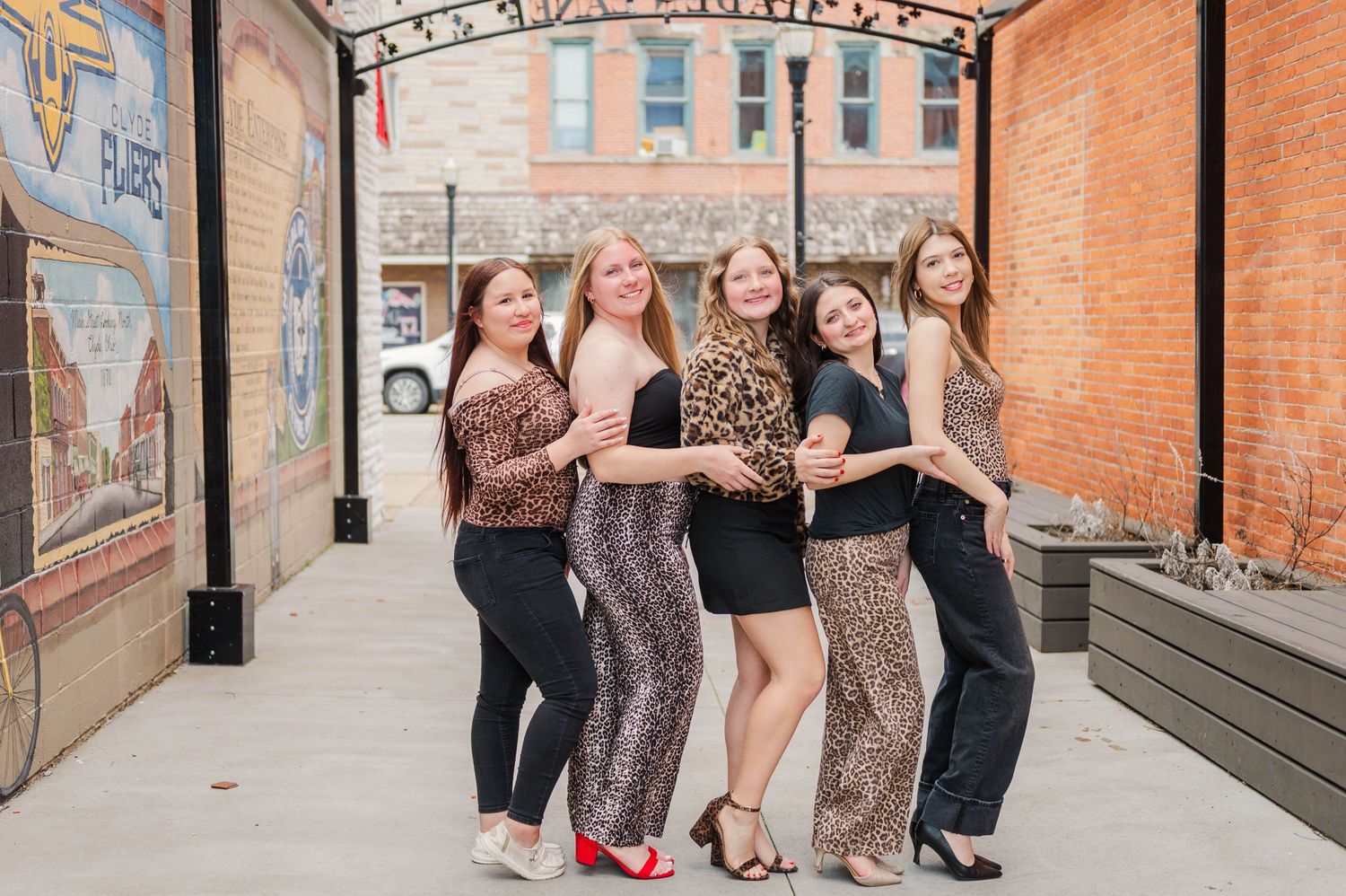 Five women posing together outdoors near a brick wall mural, wearing stylish outfits including leopard print dresses.