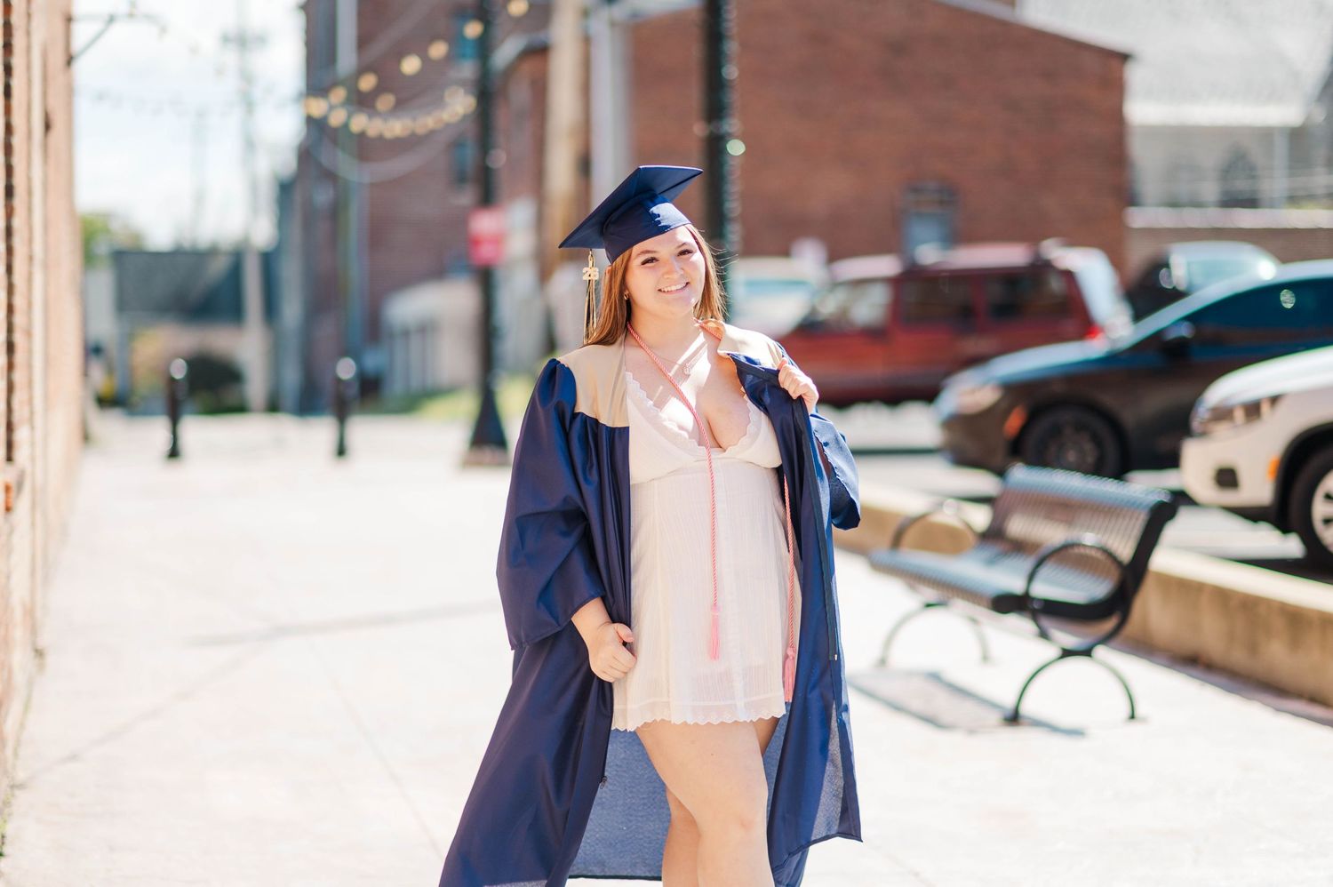 Smiling graduate in navy cap and gown posing on sunny urban sidewalk.