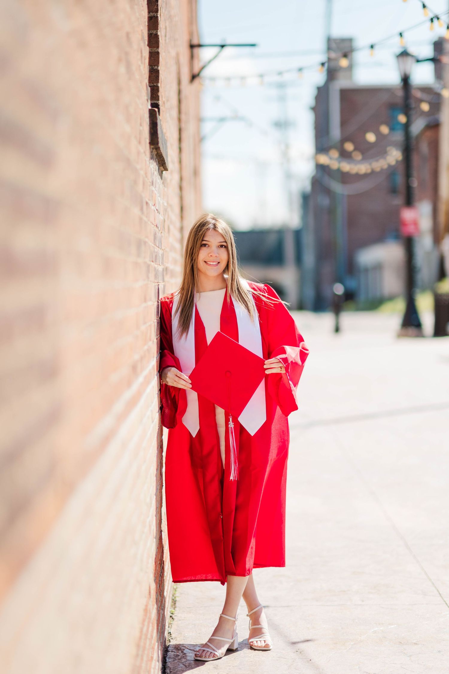 Young woman in red graduation gown and cap leaning against brick wall on sunny urban sidewalk.