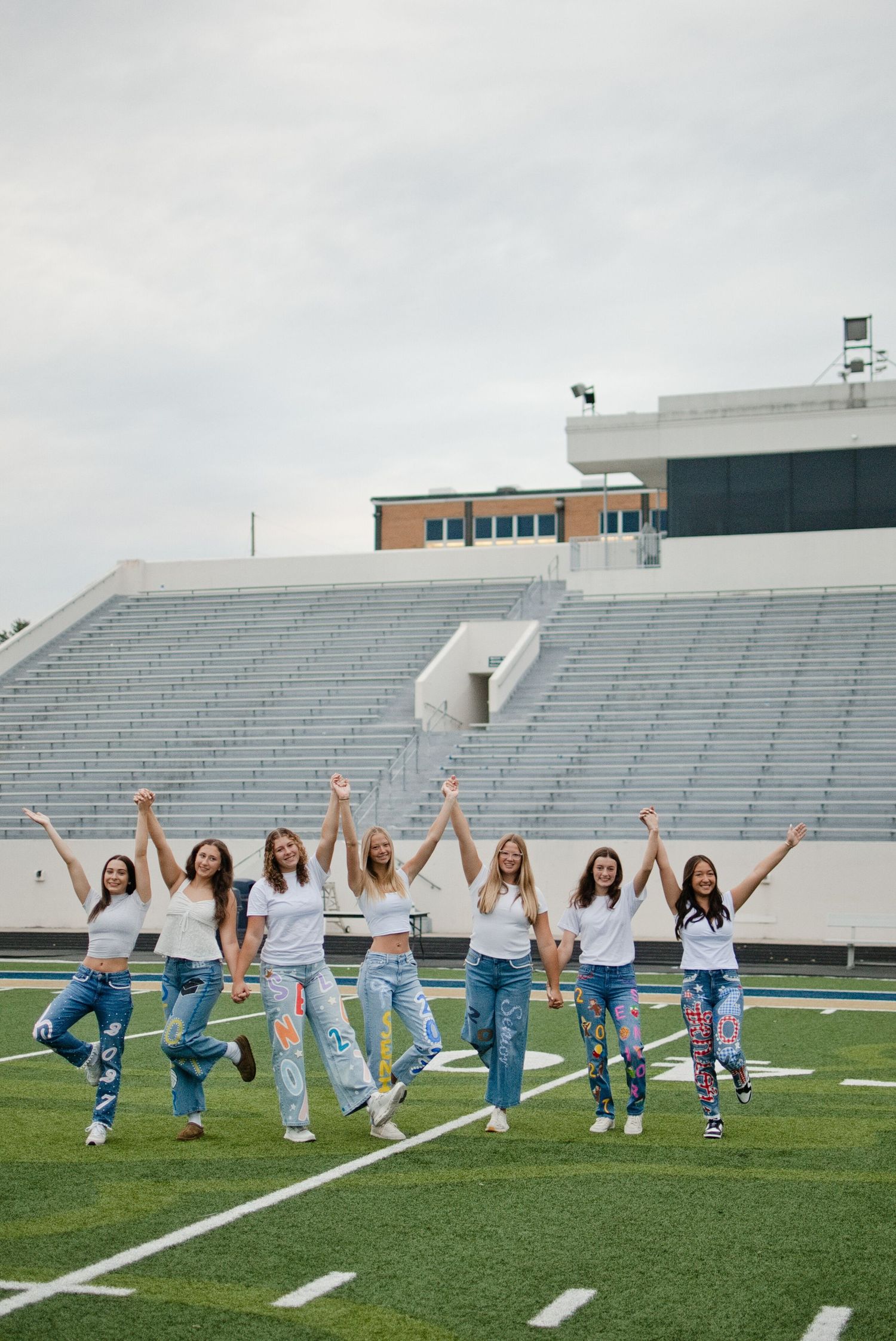 Eight girls in white tops and jeans celebrate with raised arms on a football field stadium.