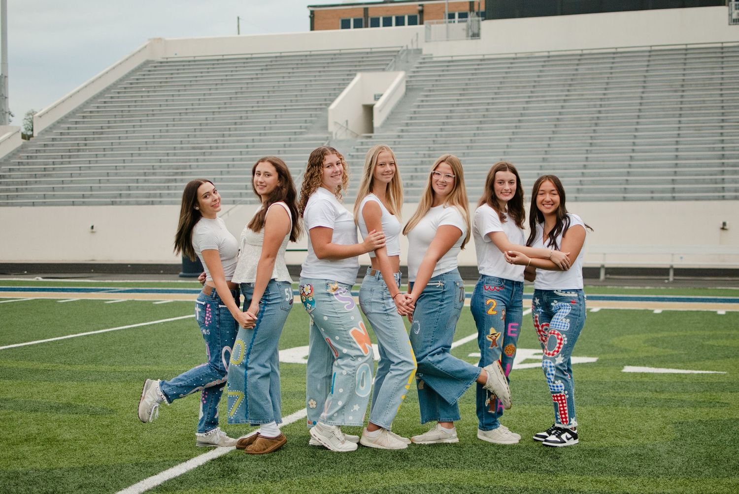 Seven young women in white tops and jeans pose together on a football field with stadium bleachers in the background.