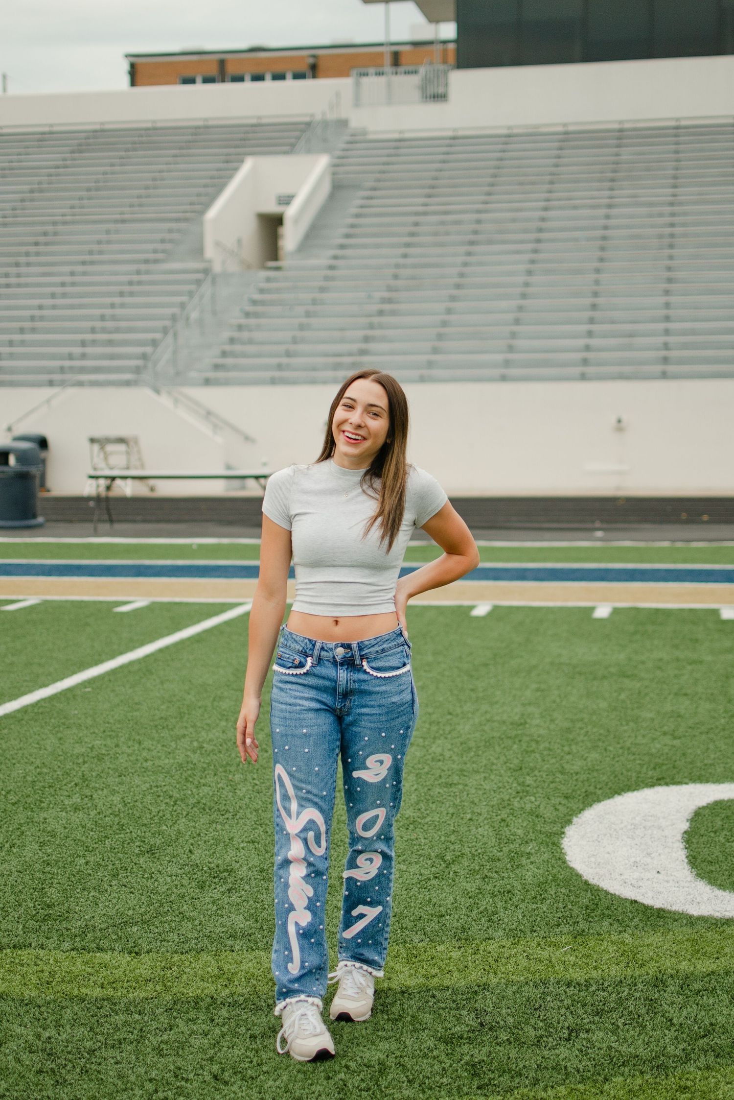 Smiling teen girl in crop top and decorated jeans poses on a football field stadium background.