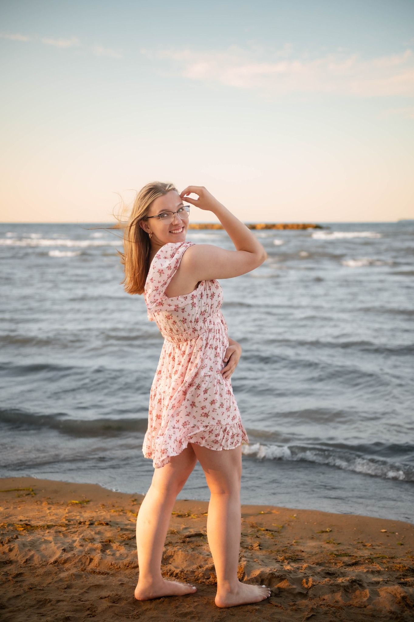A person in a floral dress poses playfully on the beach at sunset.