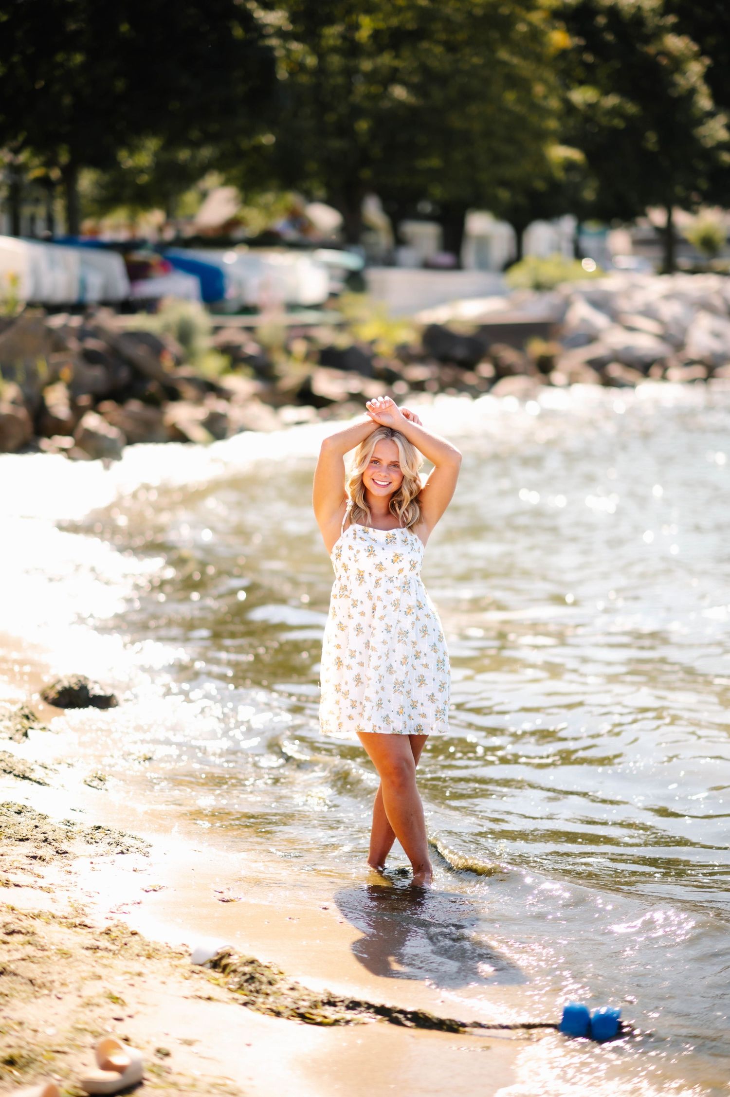 A person in a white floral dress walks through shallow ocean waves on a sunny beach day.