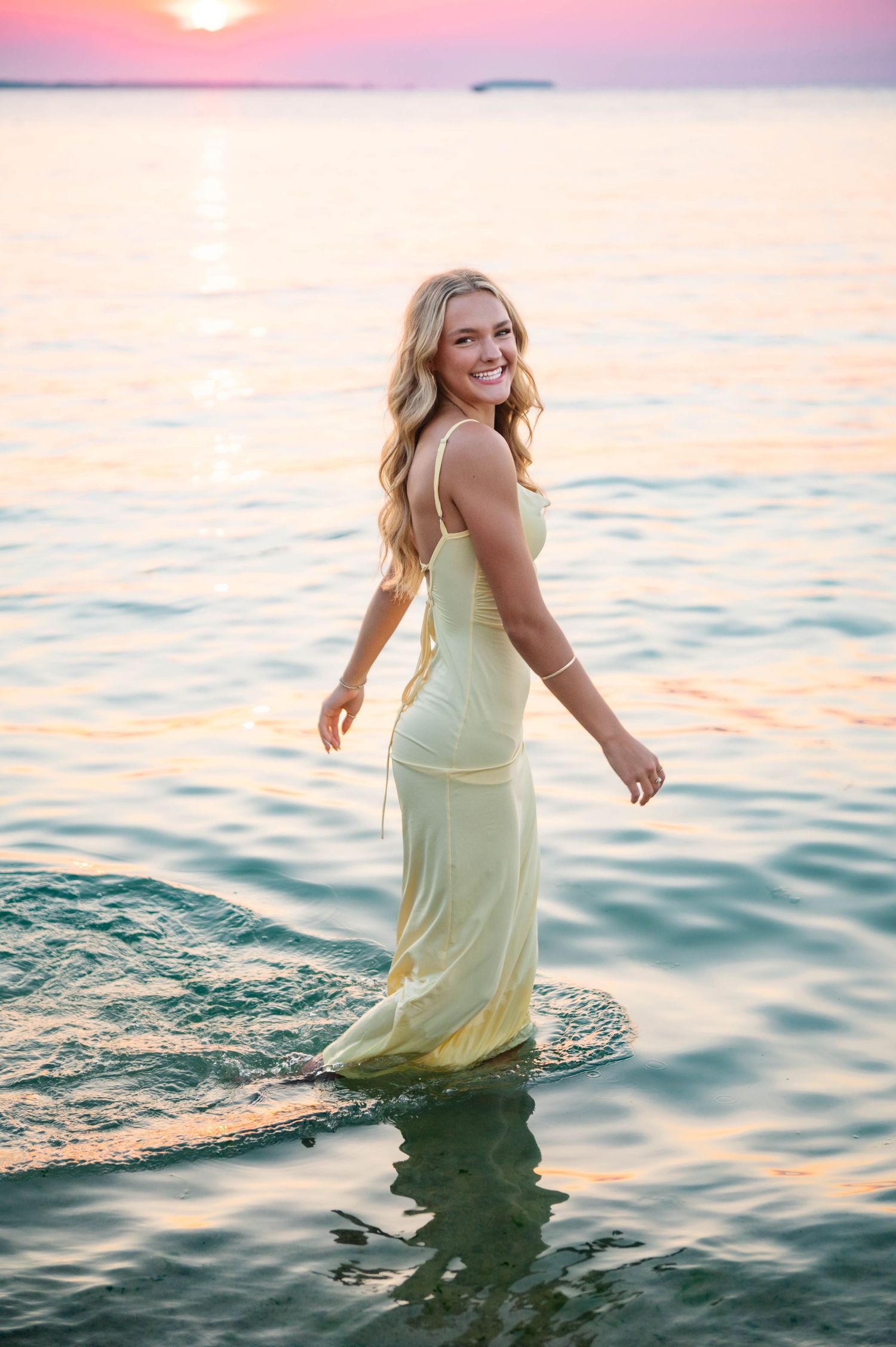 Figure in a pale yellow dress wades through ocean water during a dreamy sunset photoshoot.