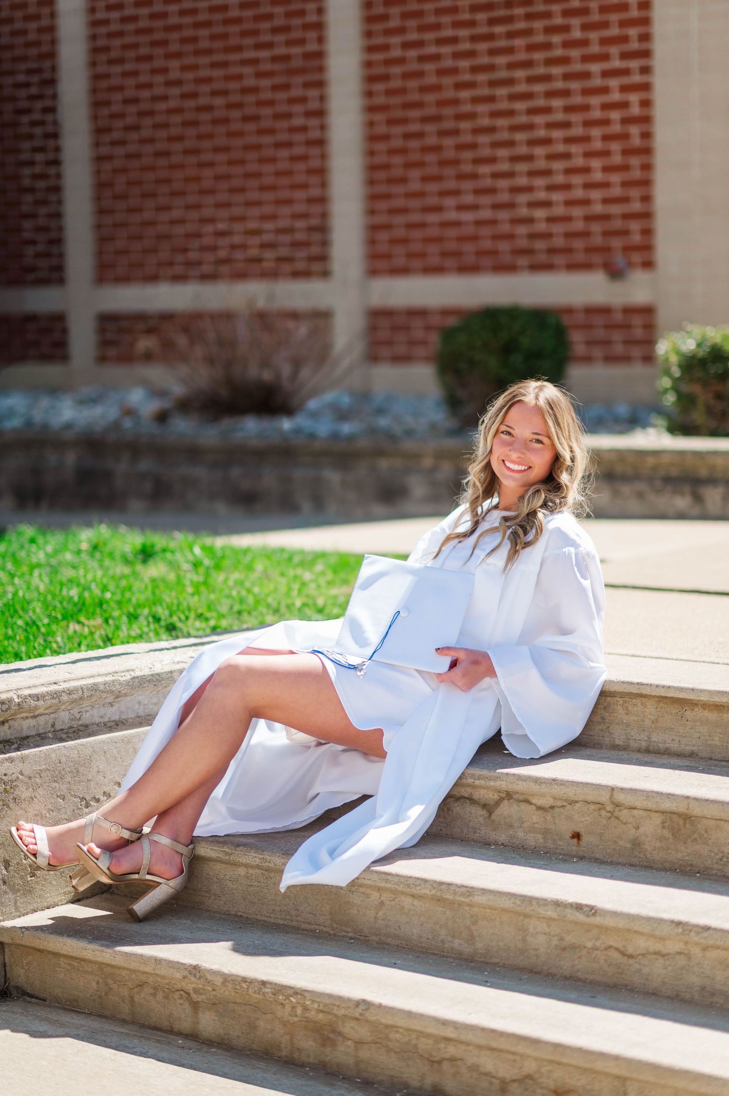 White graduation dress photoshoot on concrete steps against brick building backdrop.