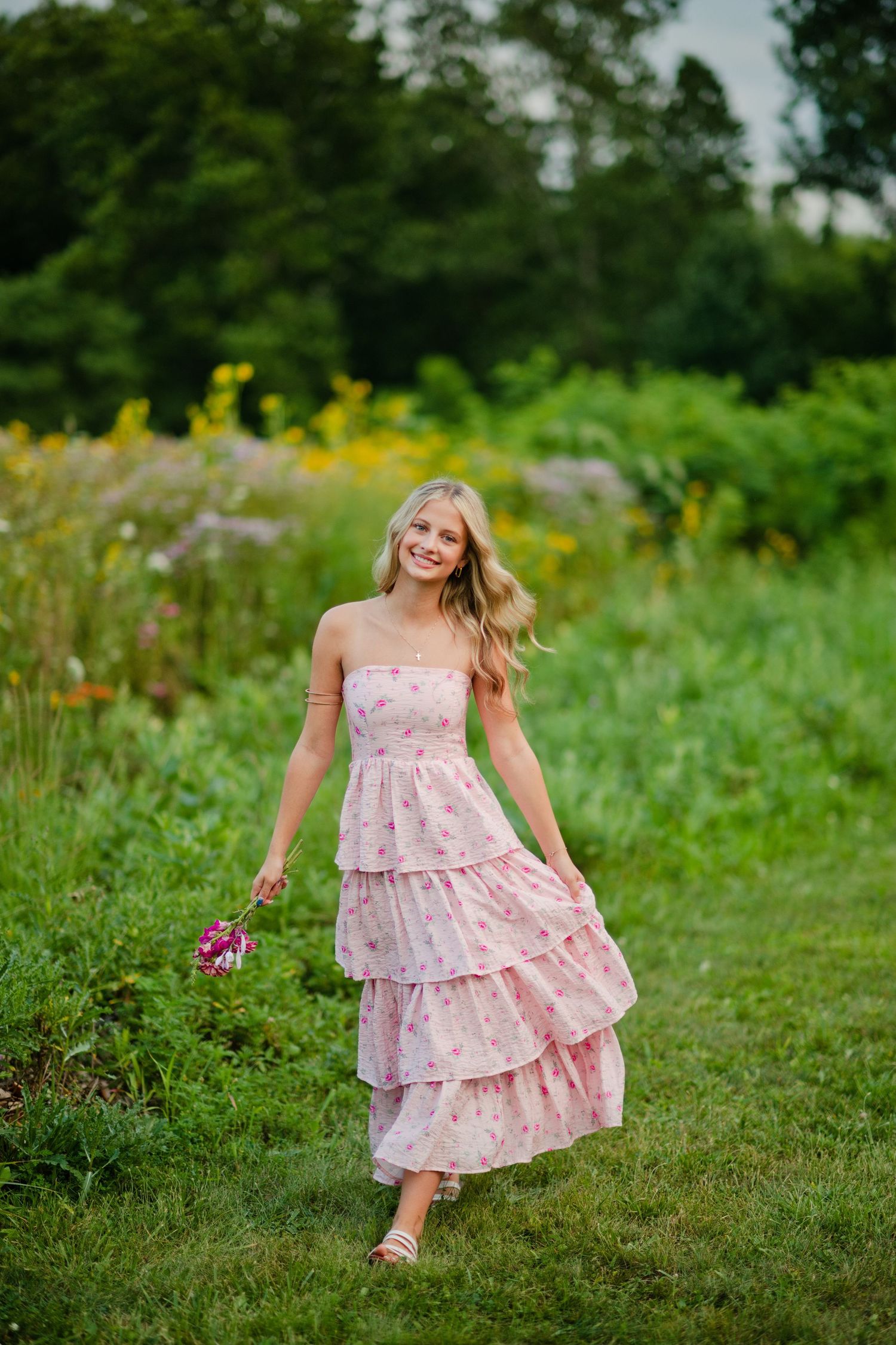 A smiling blonde woman in a pink floral tiered strapless dress holds a bouquet in a green meadow.