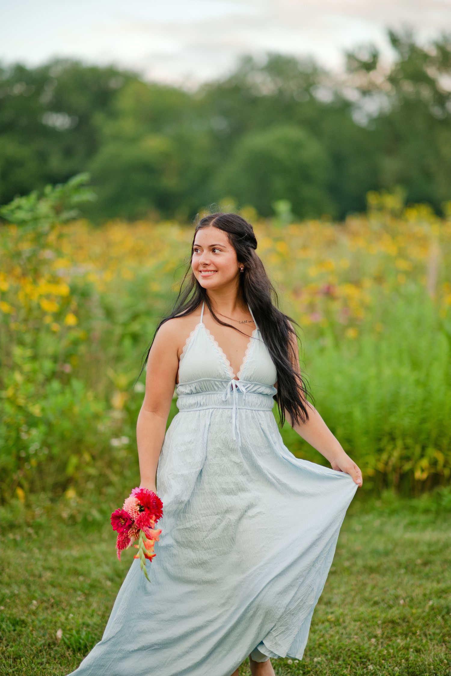 Young woman in light blue halter maxi dress smiling while holding flowers in a wildflower field.