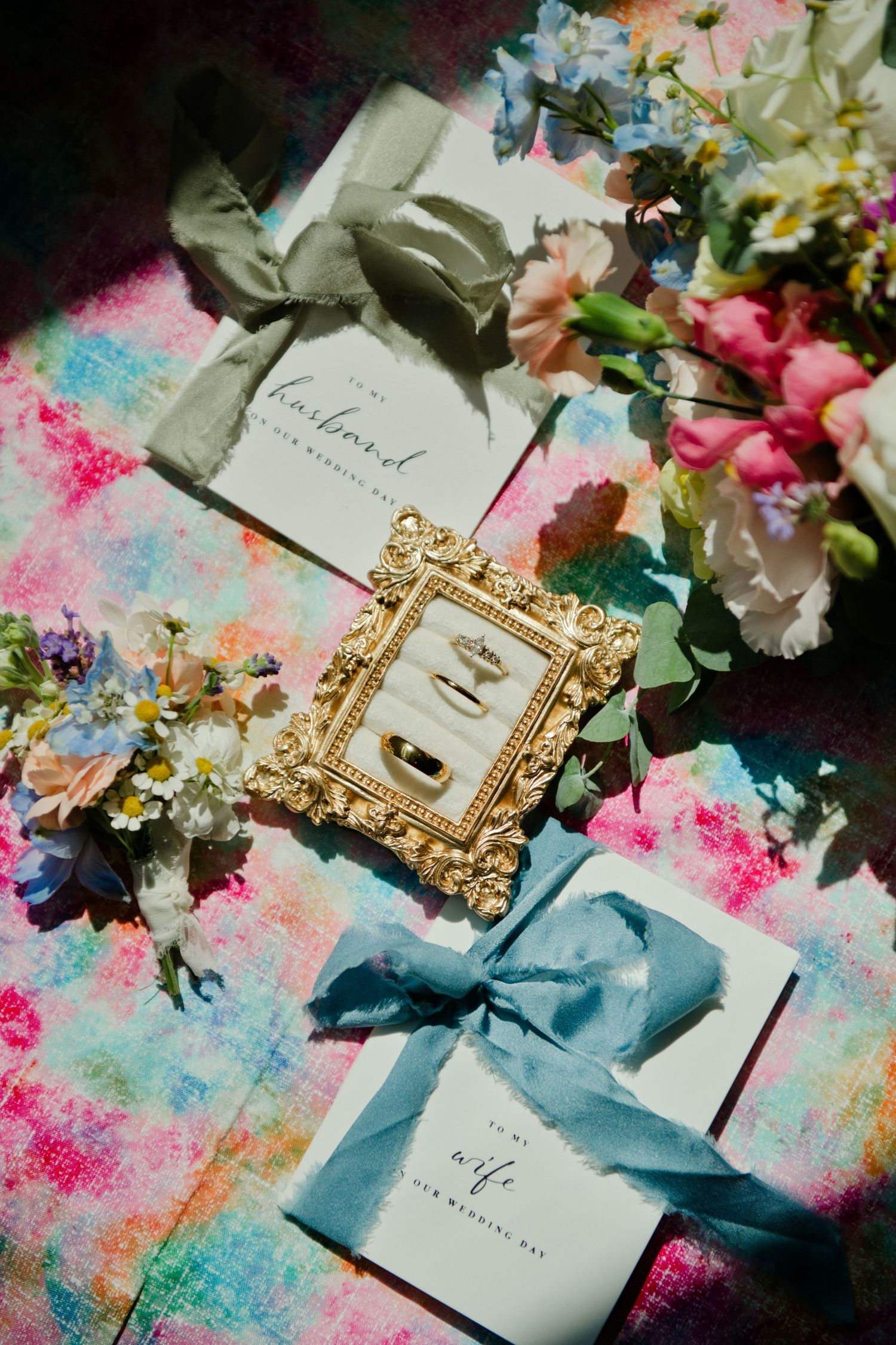 Wedding rings in gold frame with husband and wife cards, blue ribbons, and colorful floral arrangement on vibrant backdrop.