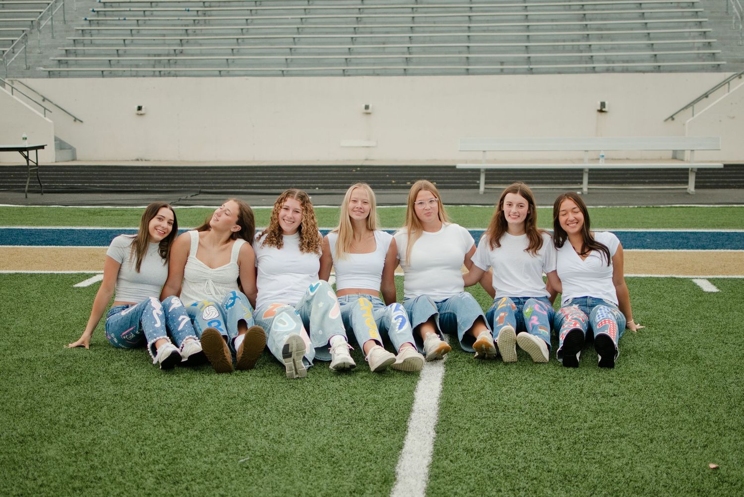 Seven young women in white tops and jeans sit together smiling on a football field with stadium bleachers behind them.