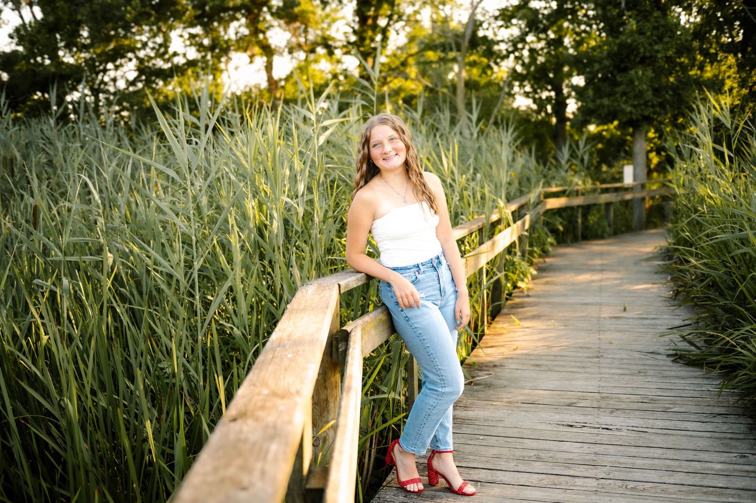 Young woman in white top and jeans poses on wooden boardwalk surrounded by tall green reeds in golden hour light.