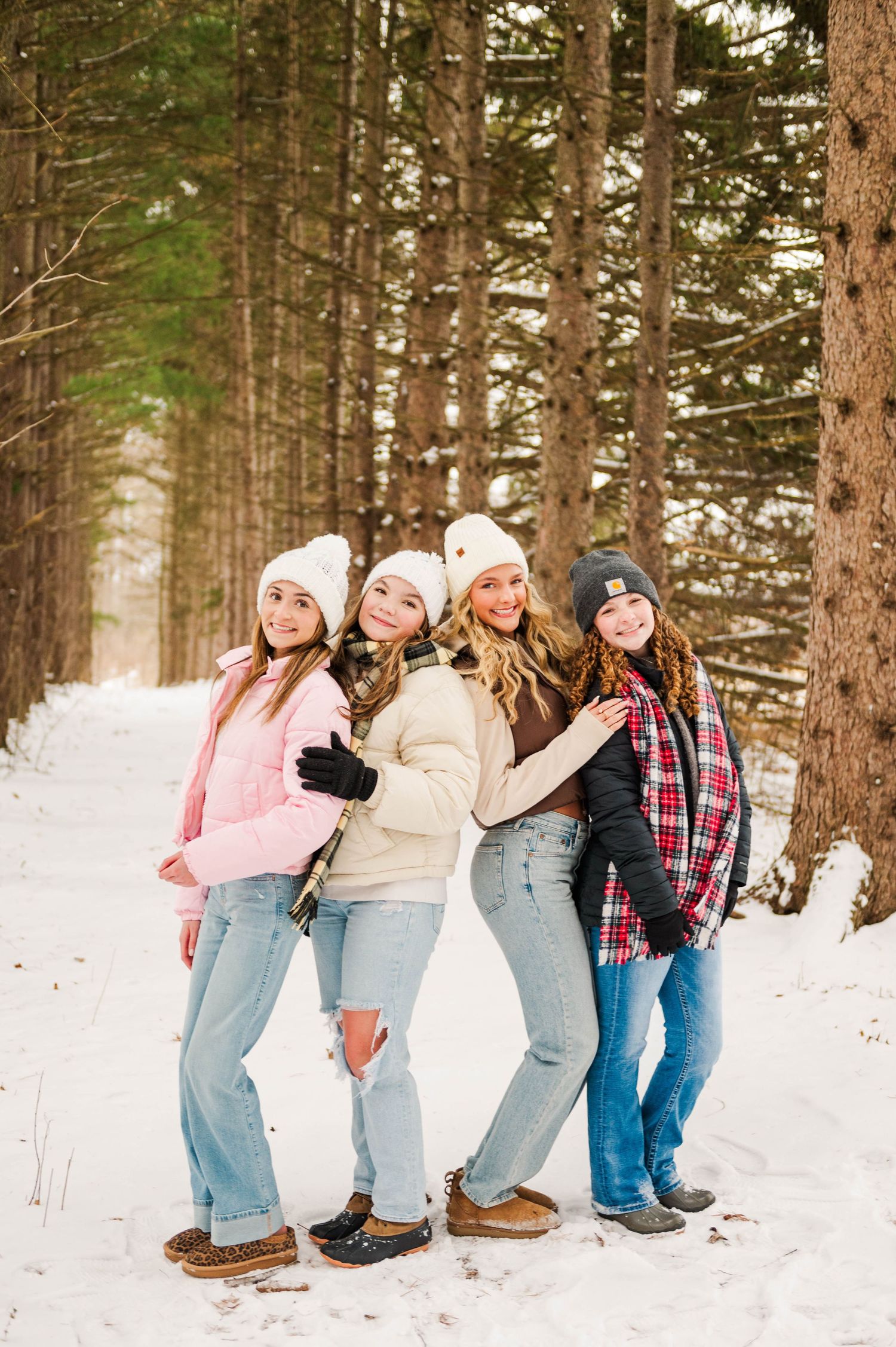 Friends in casual winter outfits stand together on a snowy path lined with tall pine trees.