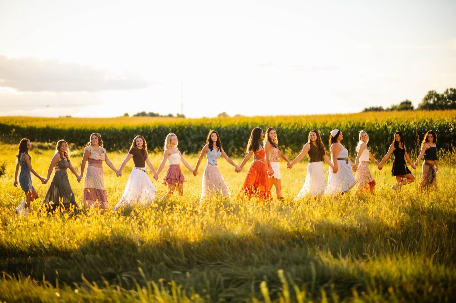 Group holding hands in a line across a sunlit field wearing summer dresses.