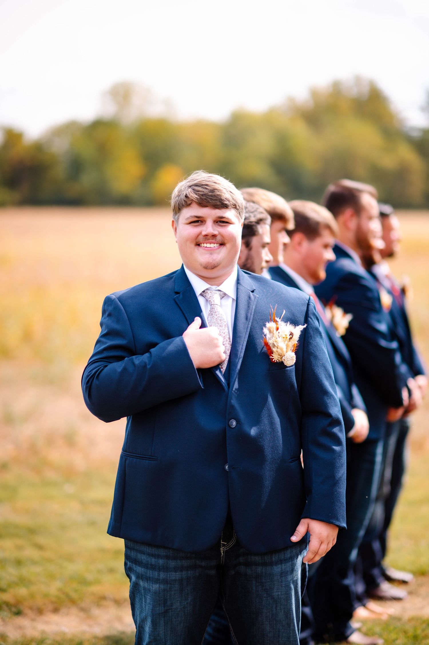Smiling groomsman in navy blue suit gives thumbs up at outdoor fall wedding with groomsmen lined up behind him.