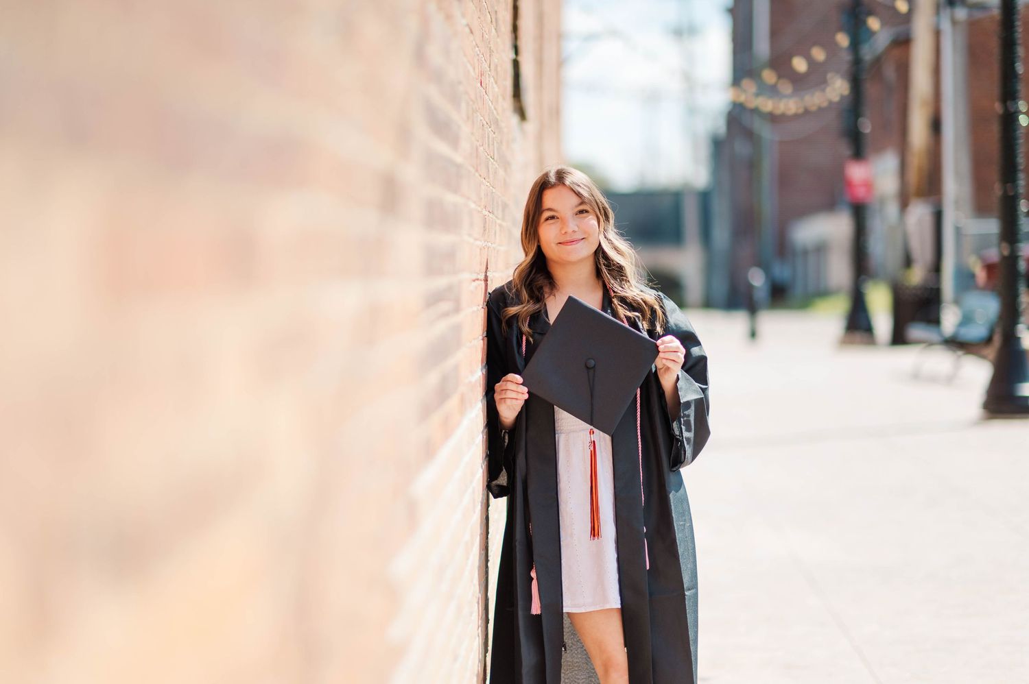 Graduate in black gown leans against a brick wall holding her cap on a sunny sidewalk.