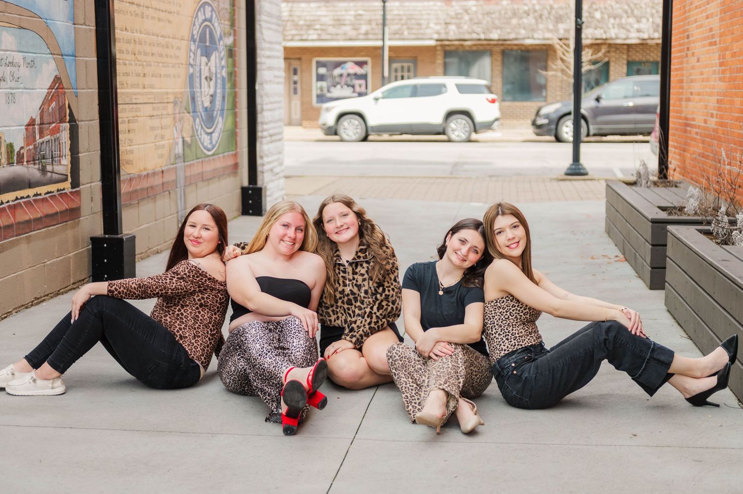 Five women smiling and posing together on a sidewalk in front of a brick building.