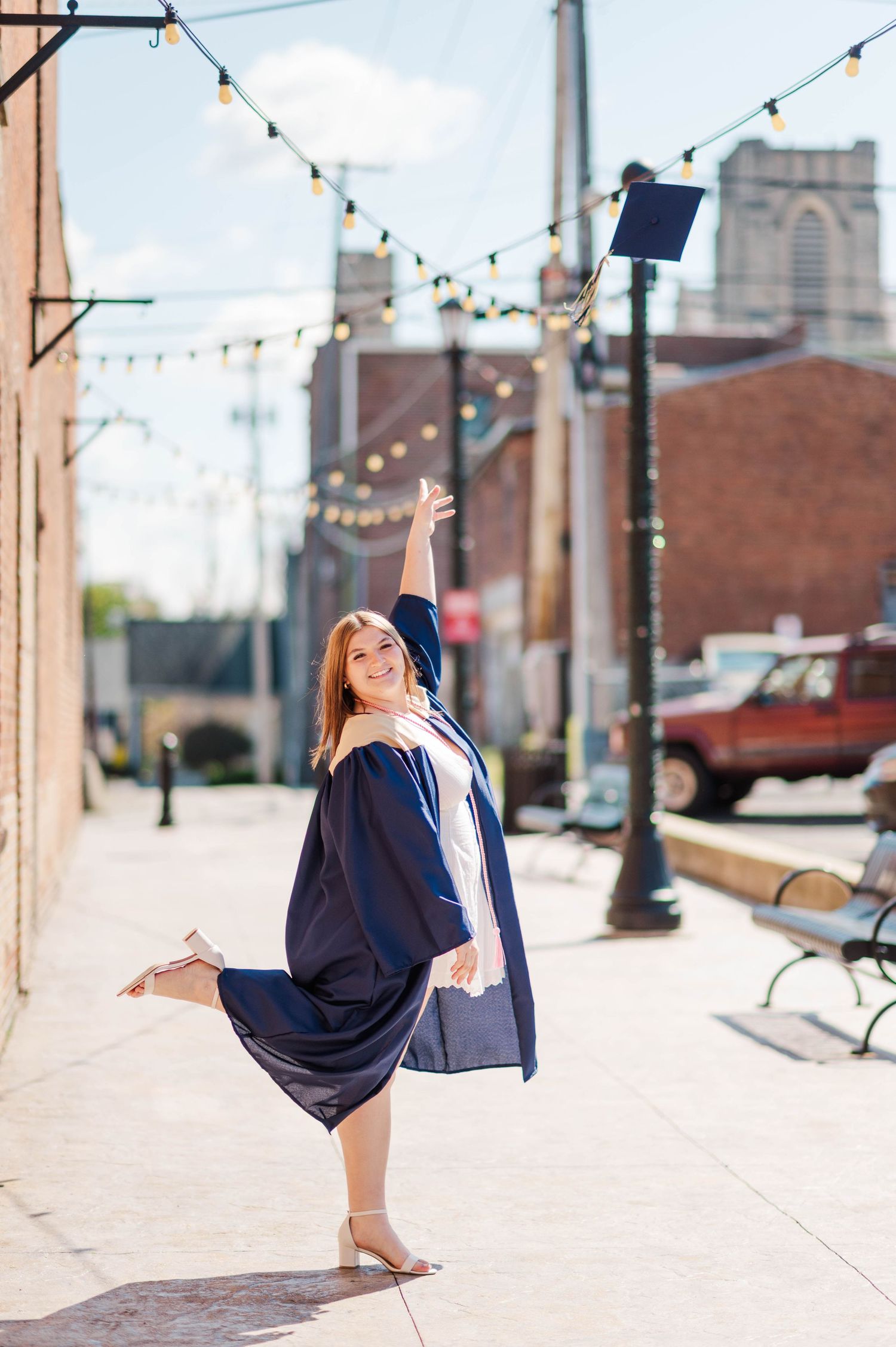 A joyful graduate in navy cap and gown tosses her cap in the air on a sunny urban sidewalk.