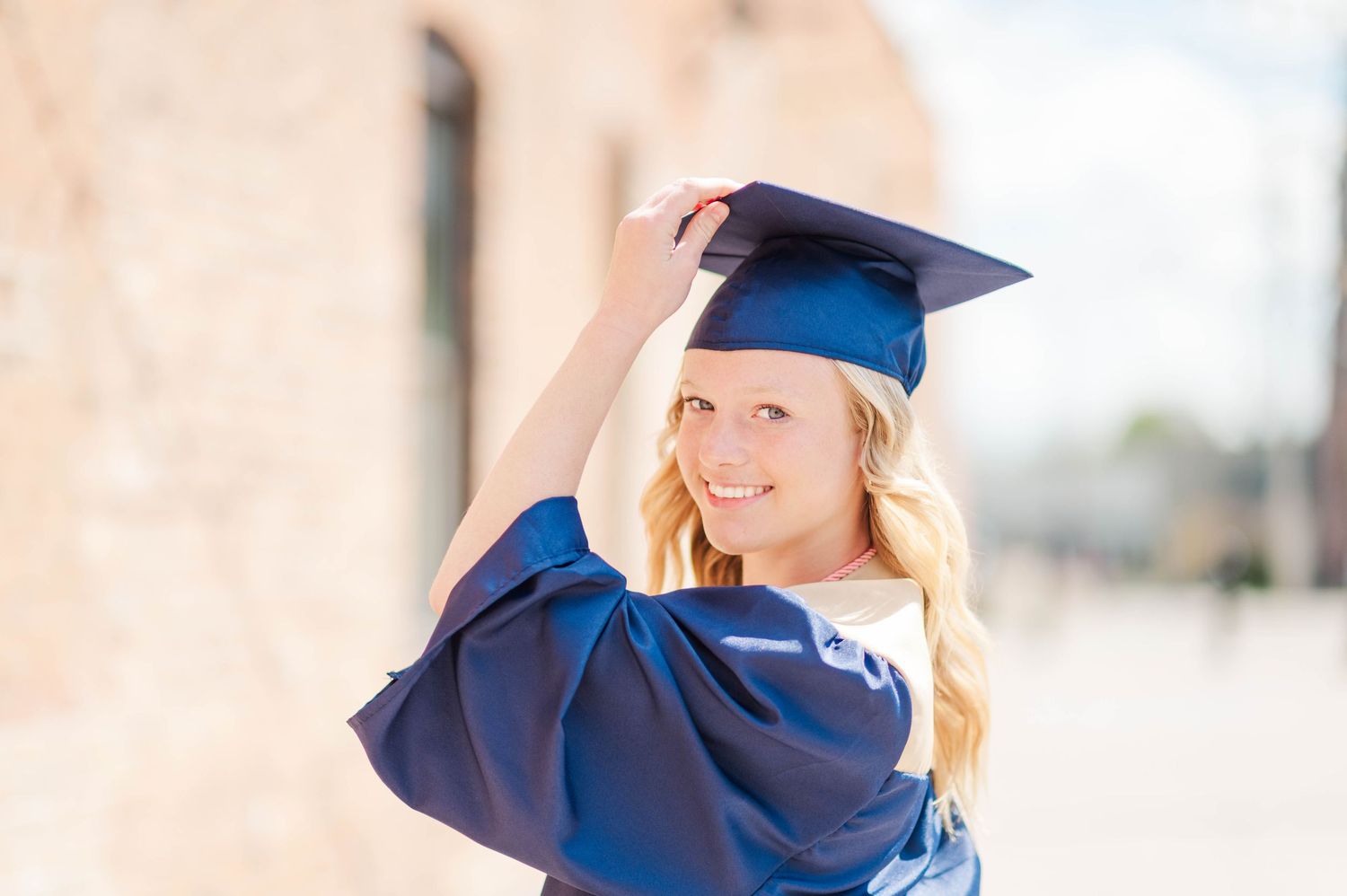 Smiling blonde graduate in navy blue cap and gown adjusting mortarboard outdoors.