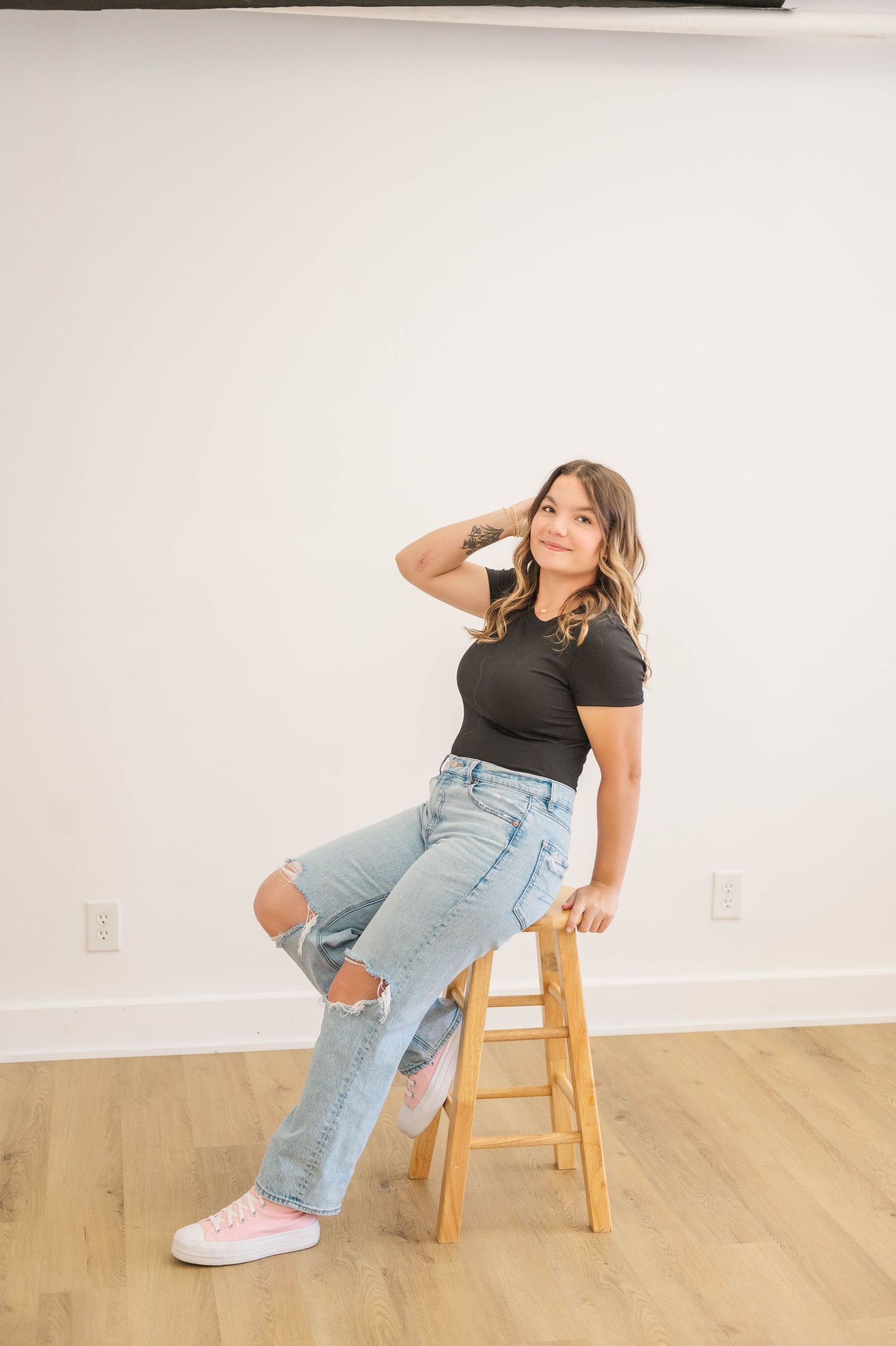 Young woman in black top and ripped jeans sits on wooden stool, hand in hair, wearing pink sneakers.